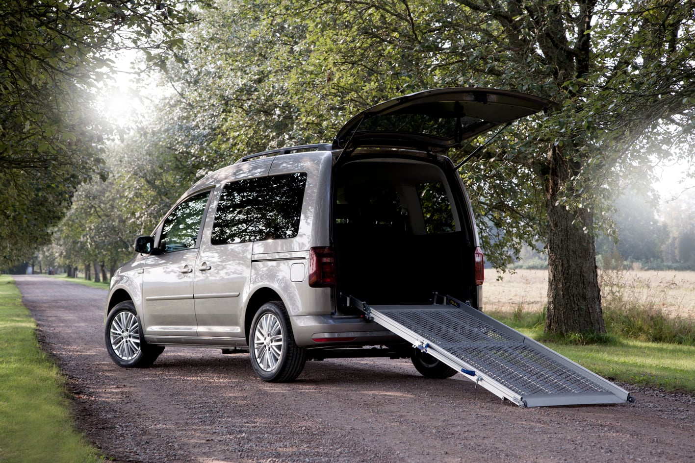 A silver minivan with its rear door open and a ramp extended, parked on a tree-lined road.