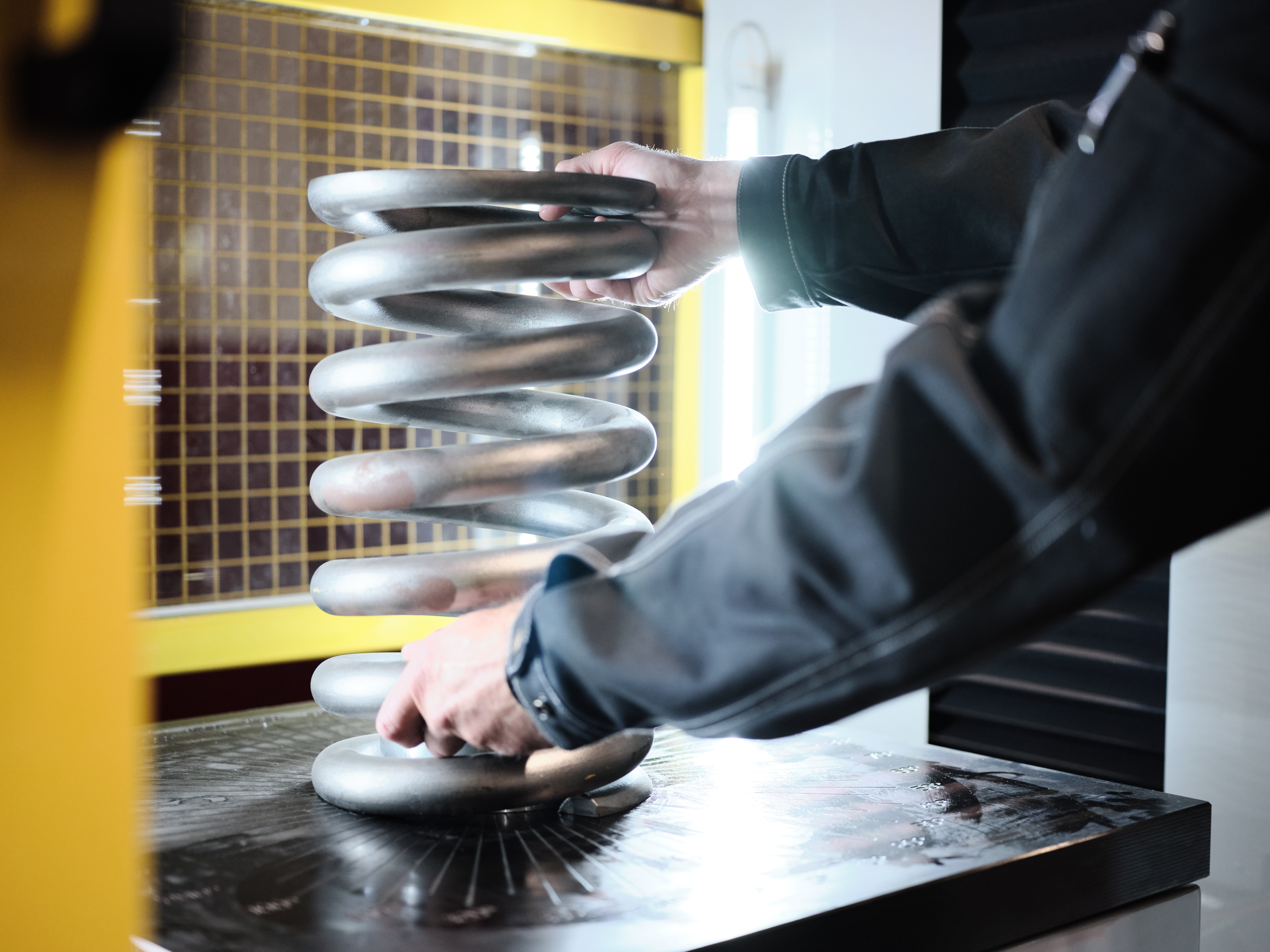 Factory worker handling a large metal coil spring on an inspection table.