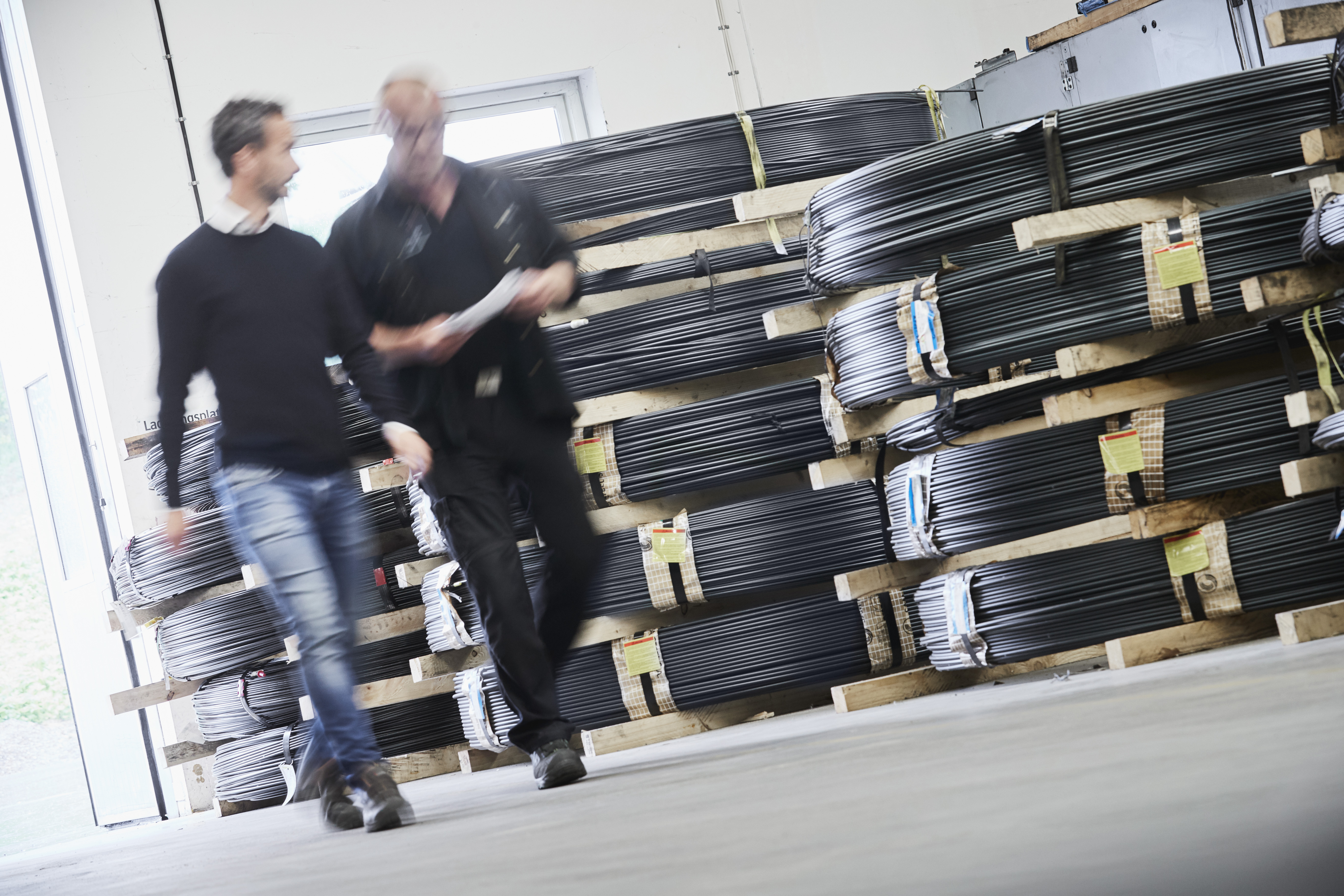 Two workers walking past stacked bundles of metal wire in a warehouse.