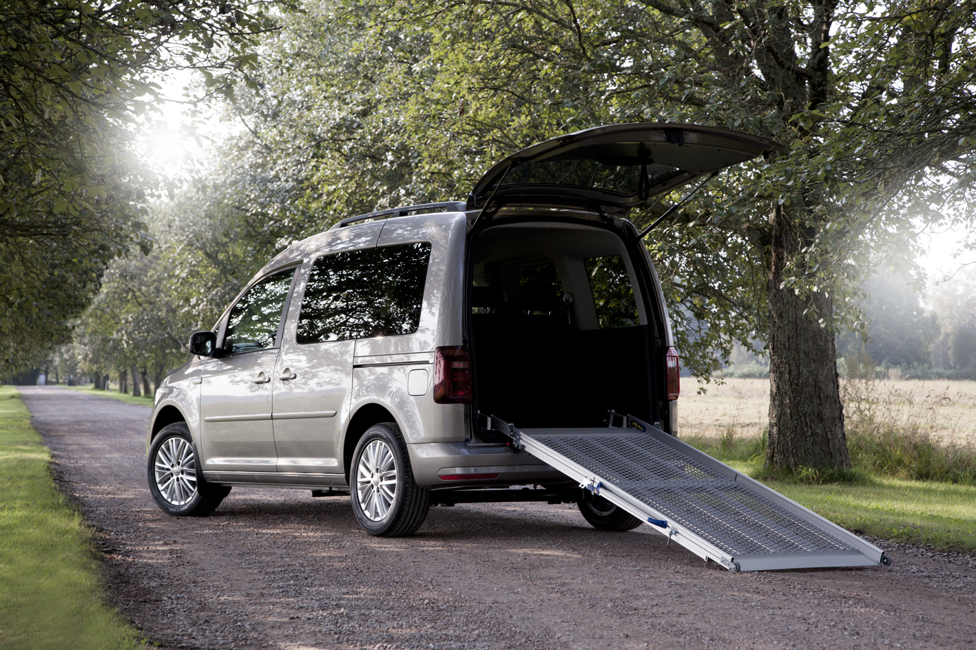A silver wheelchair-accessible van with an extended ramp is parked on a tree-lined road, conveying accessibility and independence.