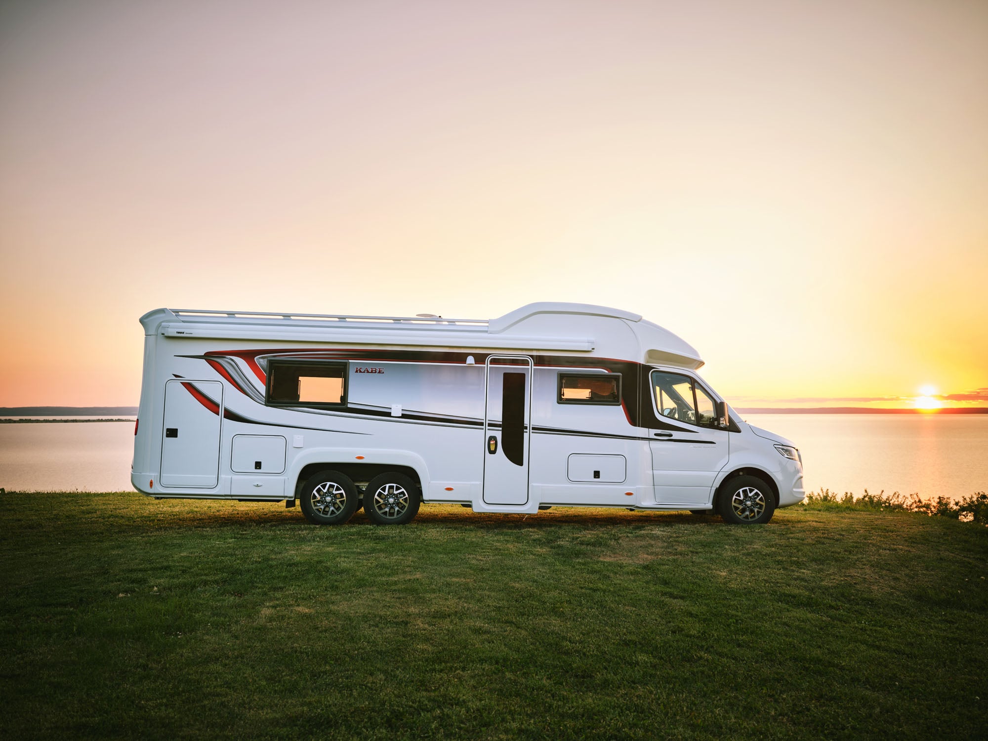 Camper van parked by a serene lakeside at sunset, with a soft orange and pink sky.