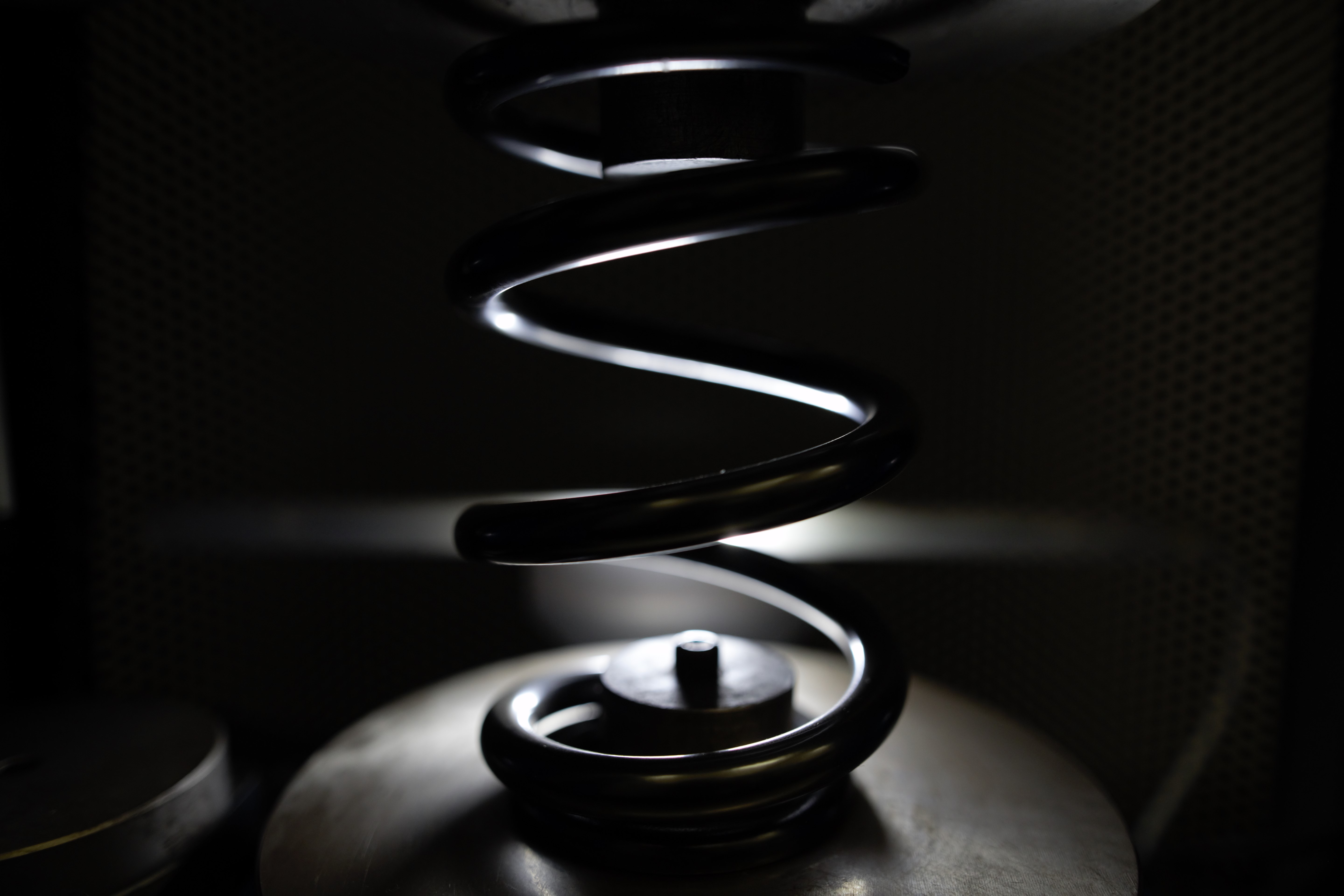 Metal coil spring under a 3D optical measuring and testing scanner, lit against a dark background.