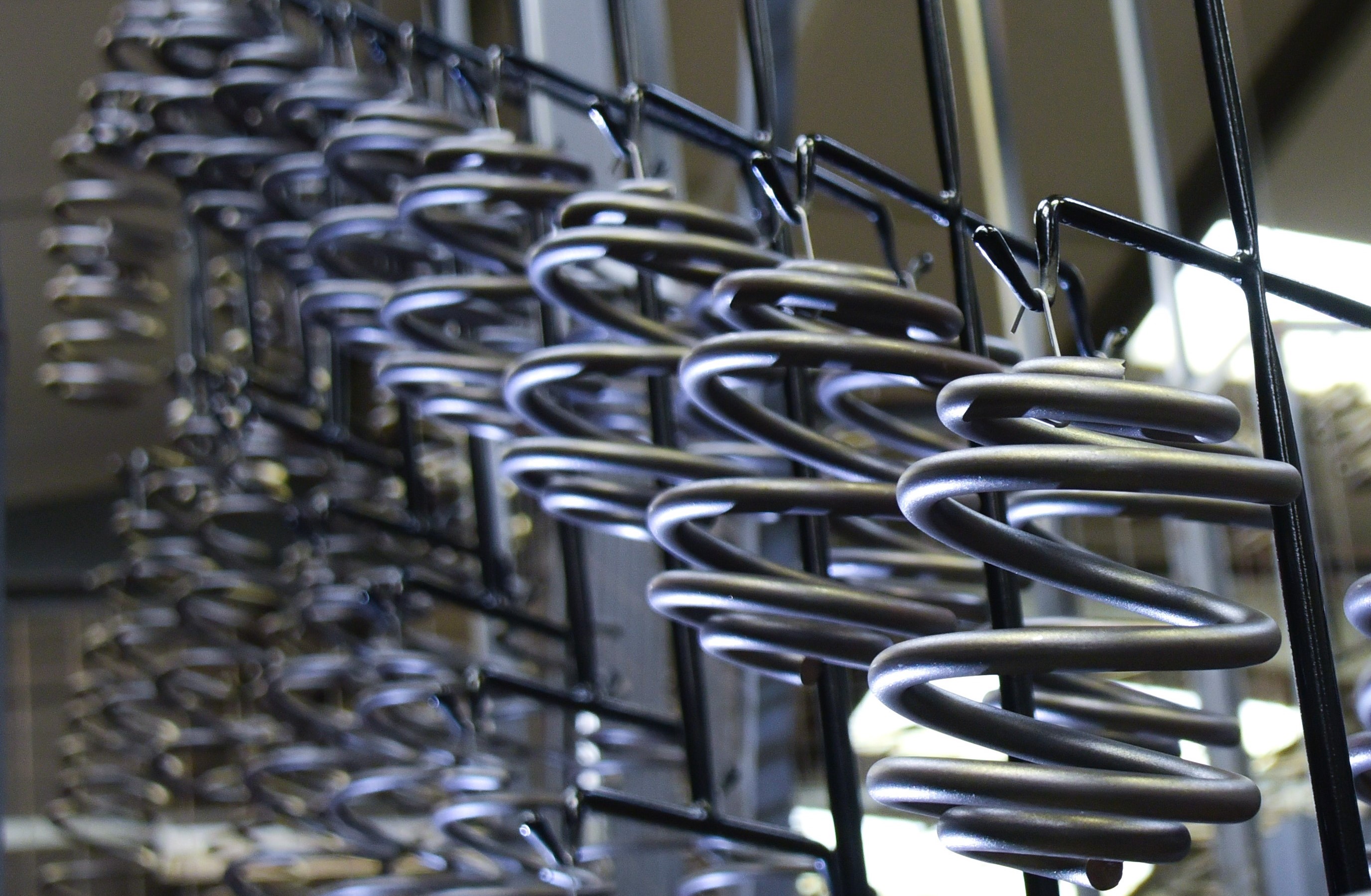 Close-up of shiny black metal coilsprings in a factory setting, with soft-focus background showing industrial machinery and a worker.