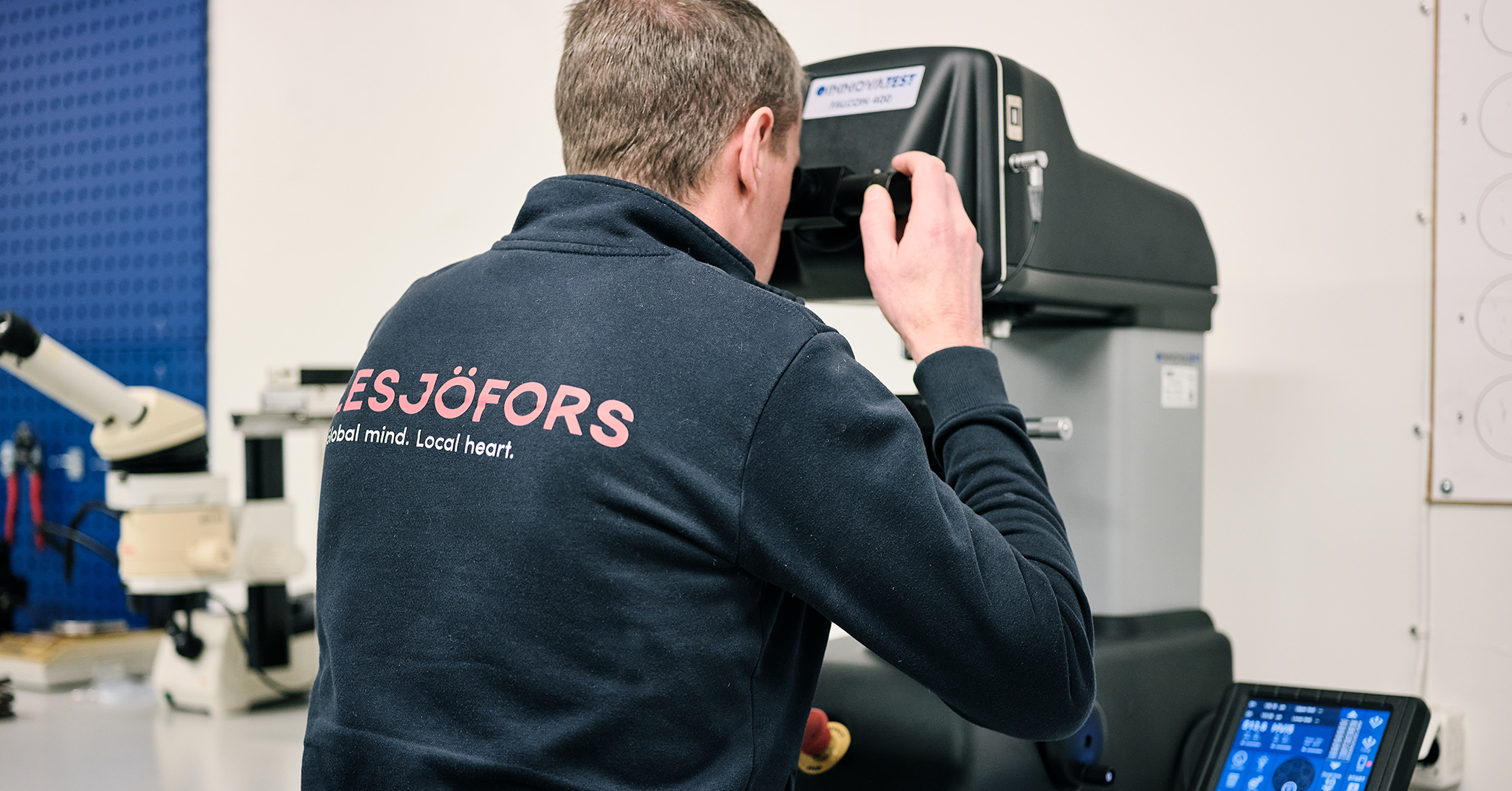 A person in a dark jacket operates a precision machine in an industrial setting, focusing intently. The environment appears technical and professional.