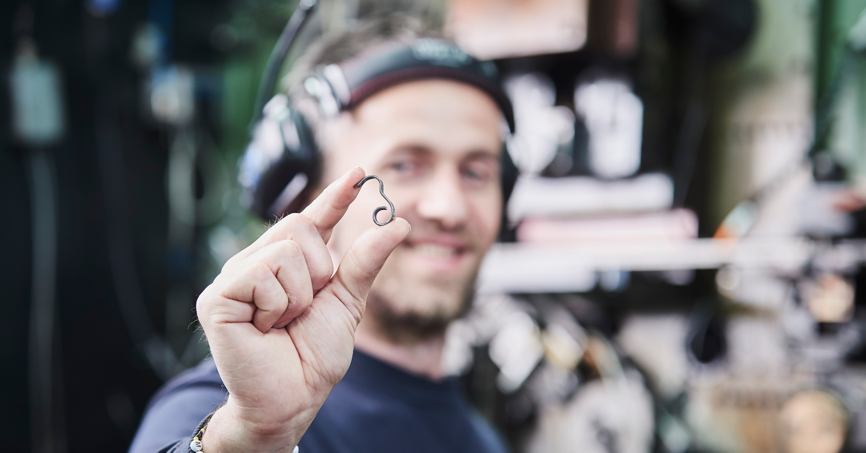 Man smiling and holding a small, shiny spring between his fingers. He wears headphones and a beanie. 