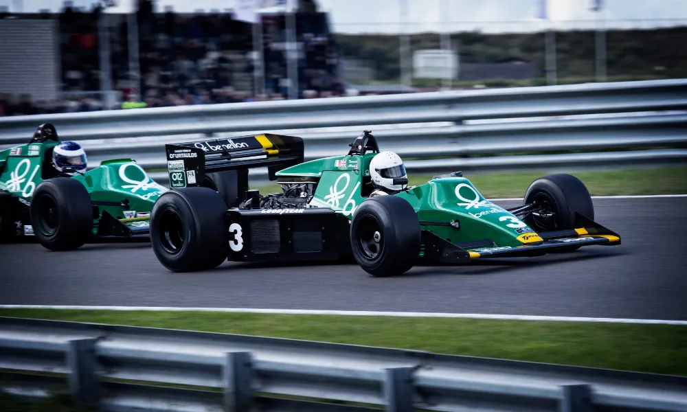 Two vintage Benetton-sponsored Formula 1 cars racing on a circuit, the lead car numbered 3 in green and black livery.