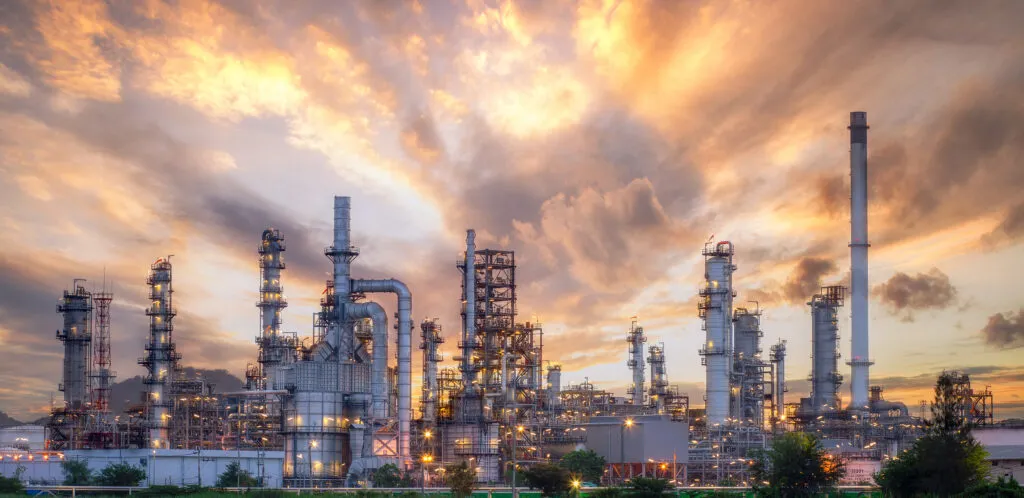 An illuminated oil refinery complex photographed at sunset, with towers, pipework and industrial structures lit up against a dramatic orange and purple sky.