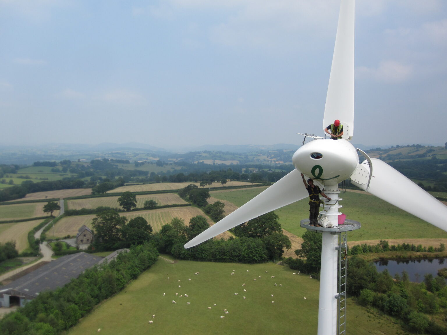 A person stands atop a large white wind turbine against a backdrop of lush, green fields and distant hills under a clear blue sky. 