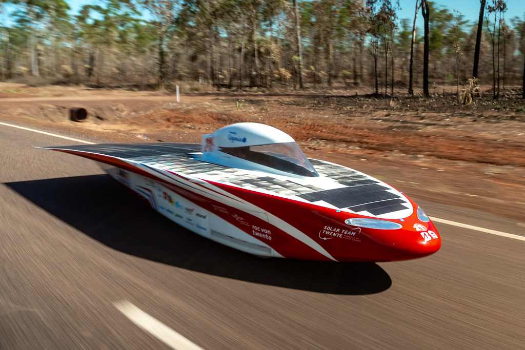 A sleek, red and white solar-powered car speeds down a rural road surrounded by trees under a clear blue sky. 