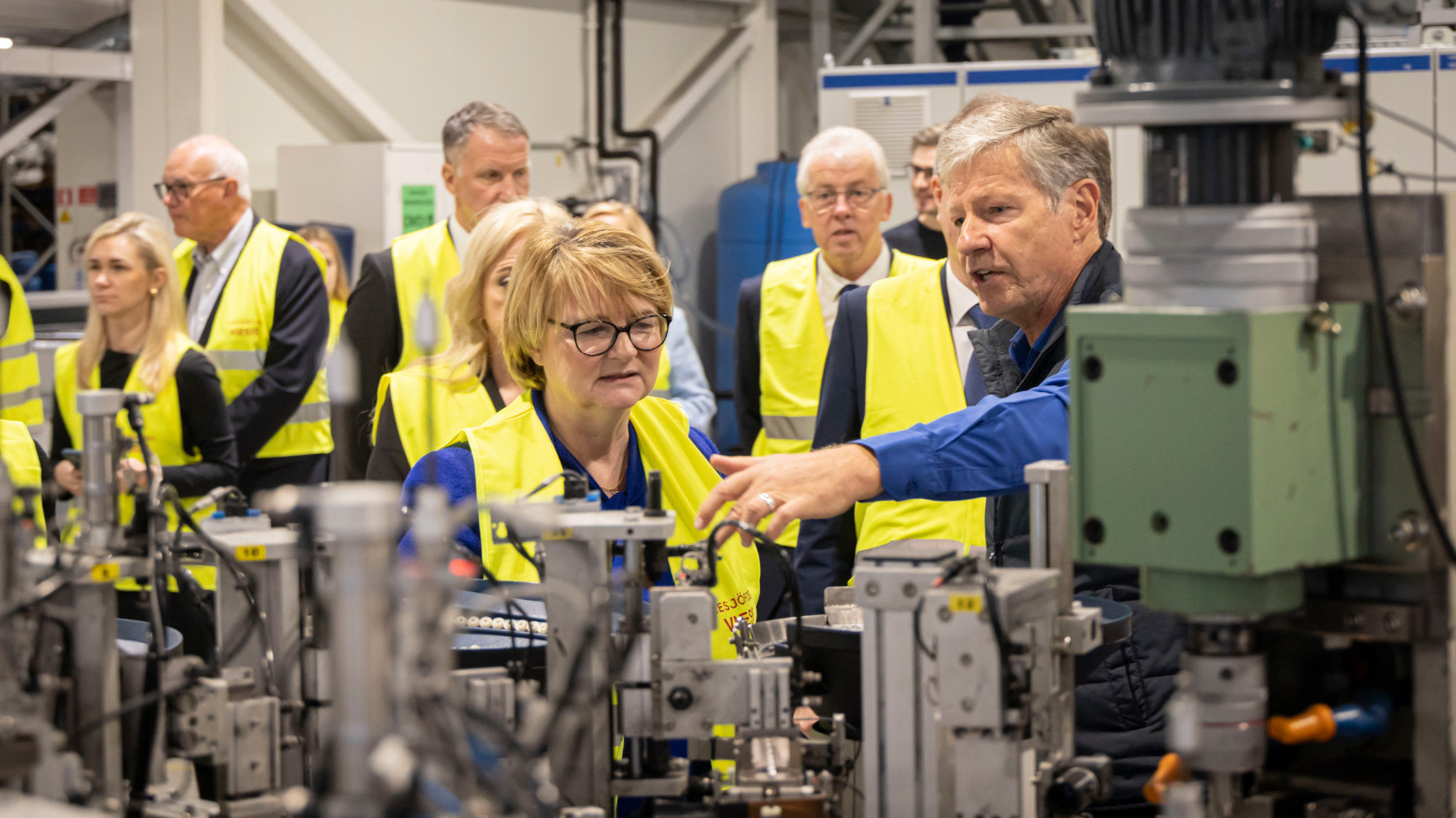 A group of people in a factory setting, wearing yellow safety vests, observe a man demonstrating machinery.
