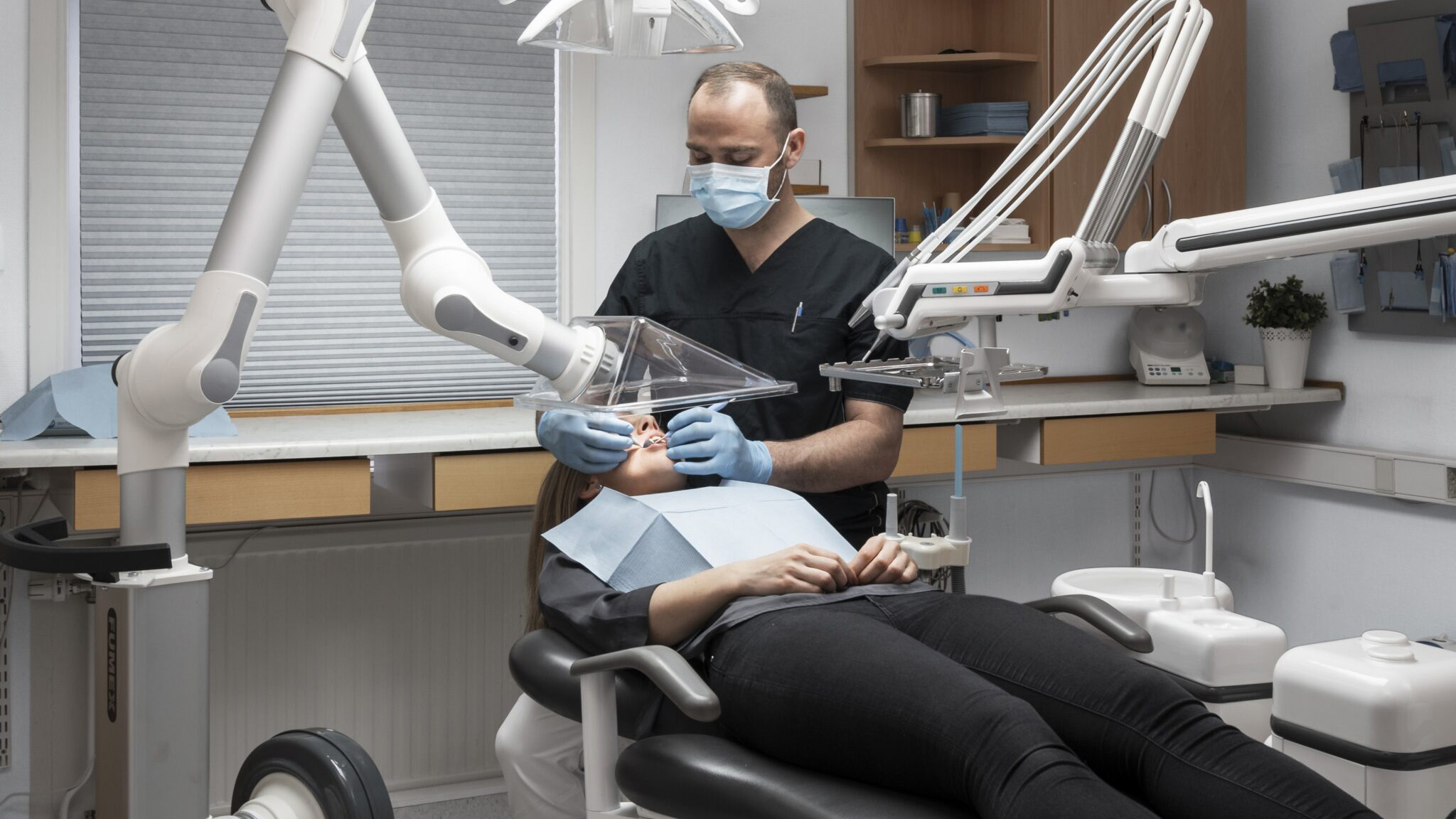 A dentist wearing a mask and gloves examines a patient's teeth under a dental light in a clean, modern clinic. The patient appears relaxed in the chair.
