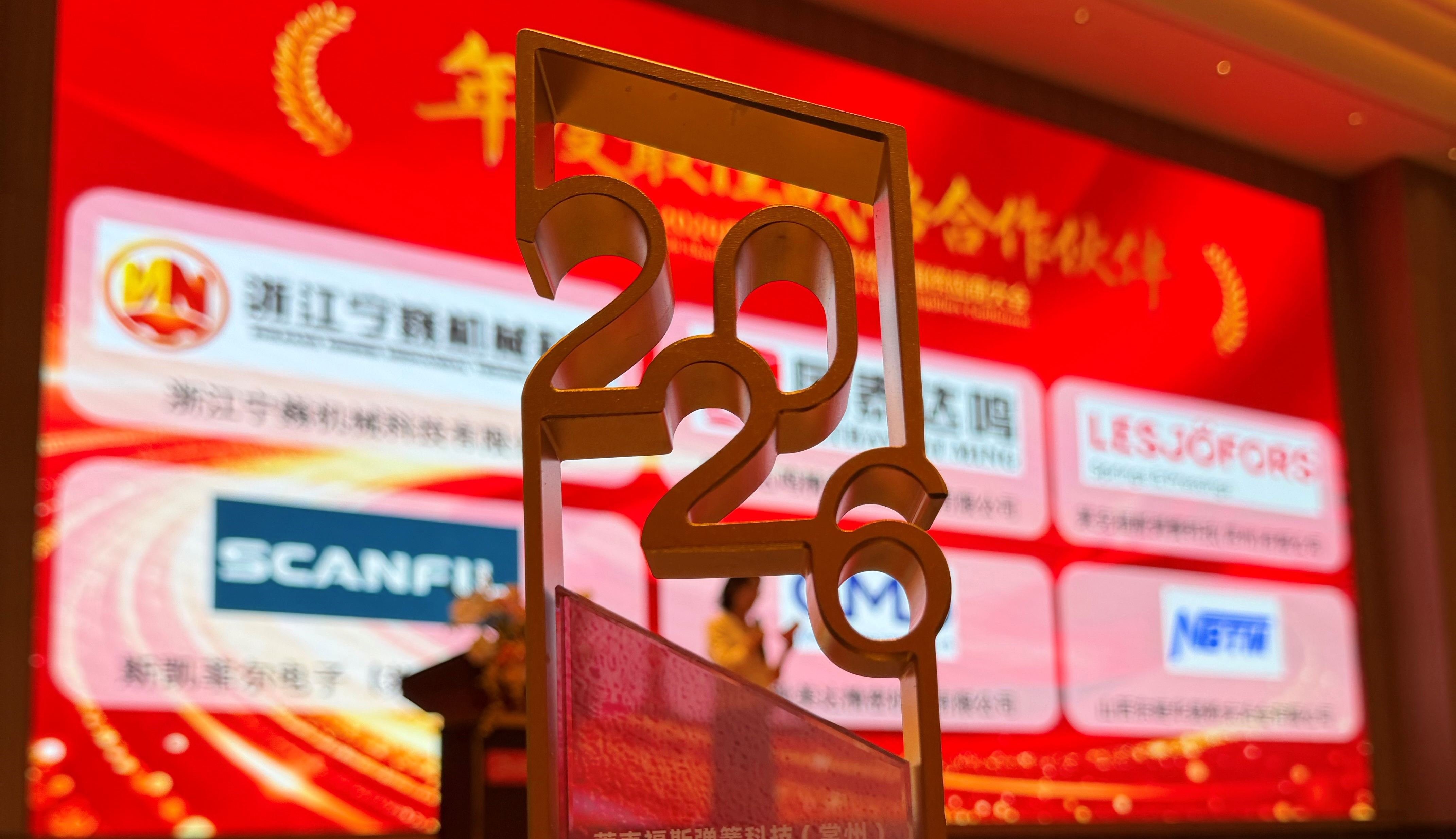 Close-up of a metallic “2022” award trophy in the foreground, with a red event backdrop featuring Chinese text, multiple company logos (including Lesjöfors and Scanfil), and a blurred speaker at a podium in the background.