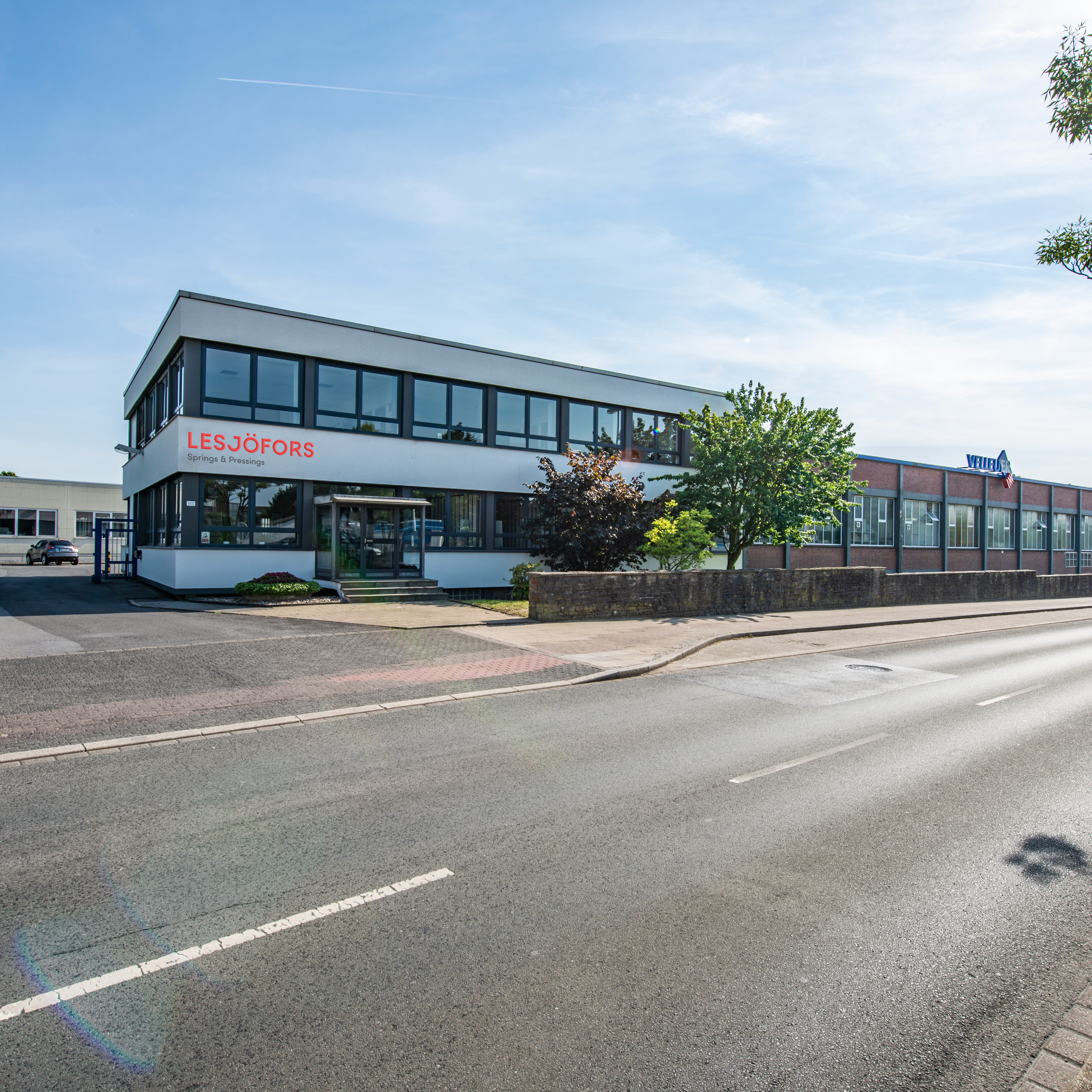 A modern industrial building labeled "Lesjöfors" with large windows sits beside a quiet road. The sky is clear, and trees line the street.