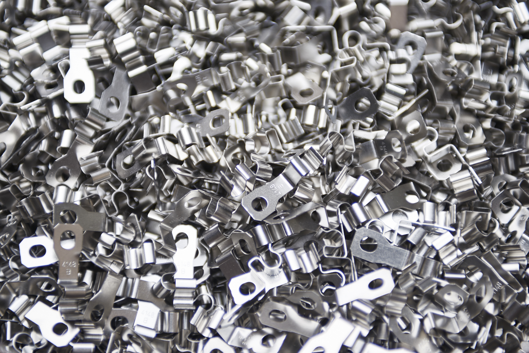 Heap of shiny, metallic stamped parts with holes, showcasing a mix of curved and flat shapes, reflecting light. Industrial and organized feel.