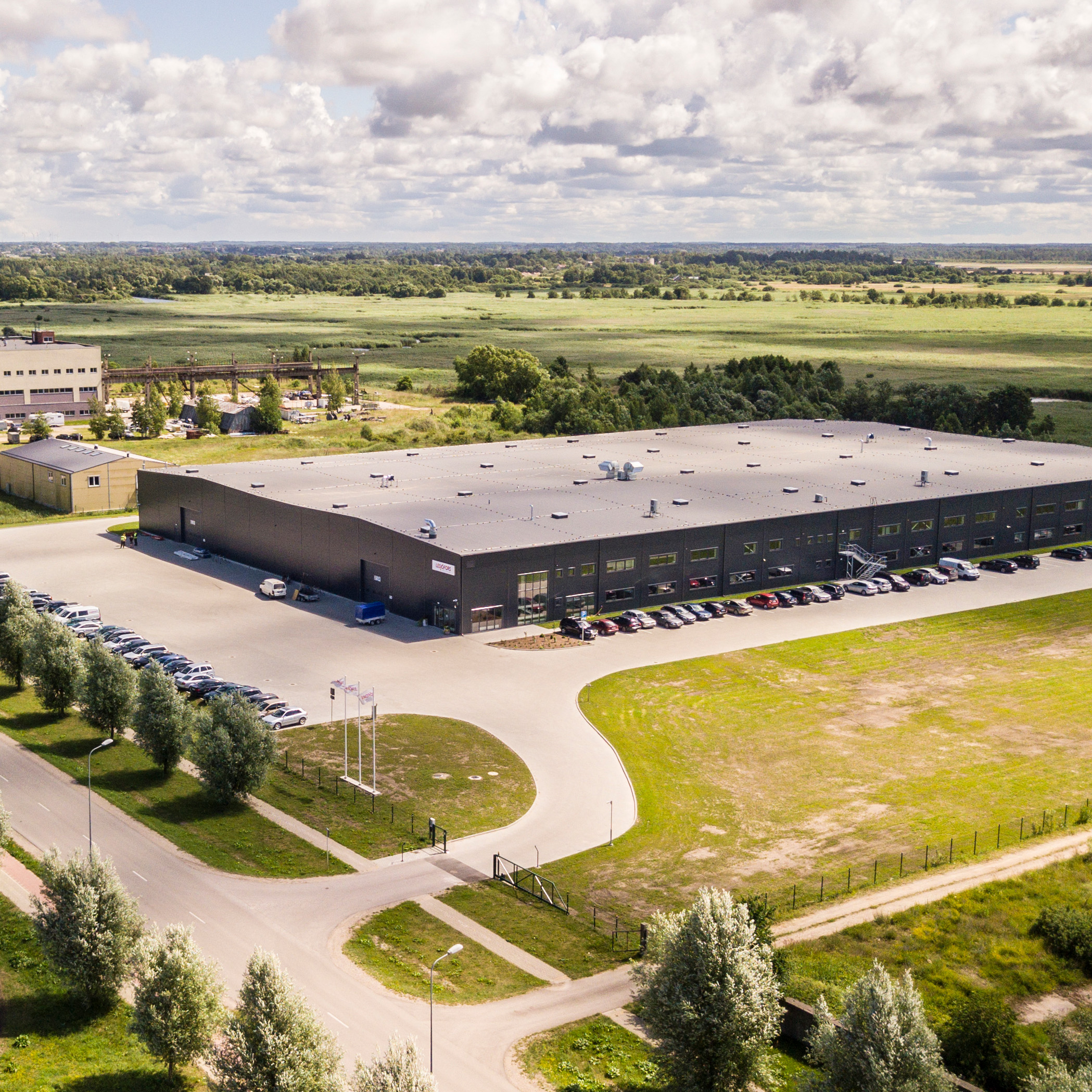 Aerial view of a large industrial building surrounded by parking lots and trees. The flat landscape extends to the horizon under a partly cloudy sky.