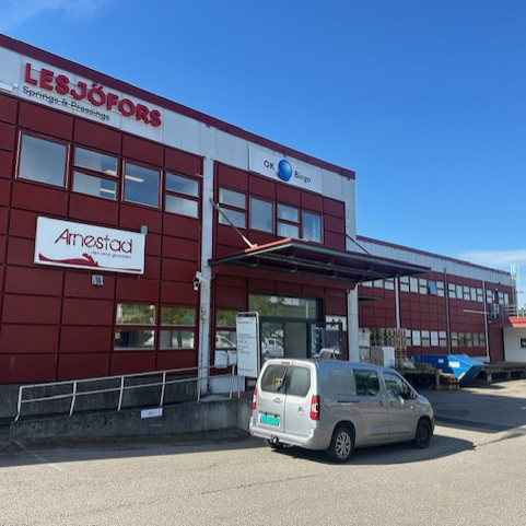 Red industrial building with "Lesjöfors," "Arnestad," and "OK Elektronik" signs. A white van is parked in front under a clear blue sky.