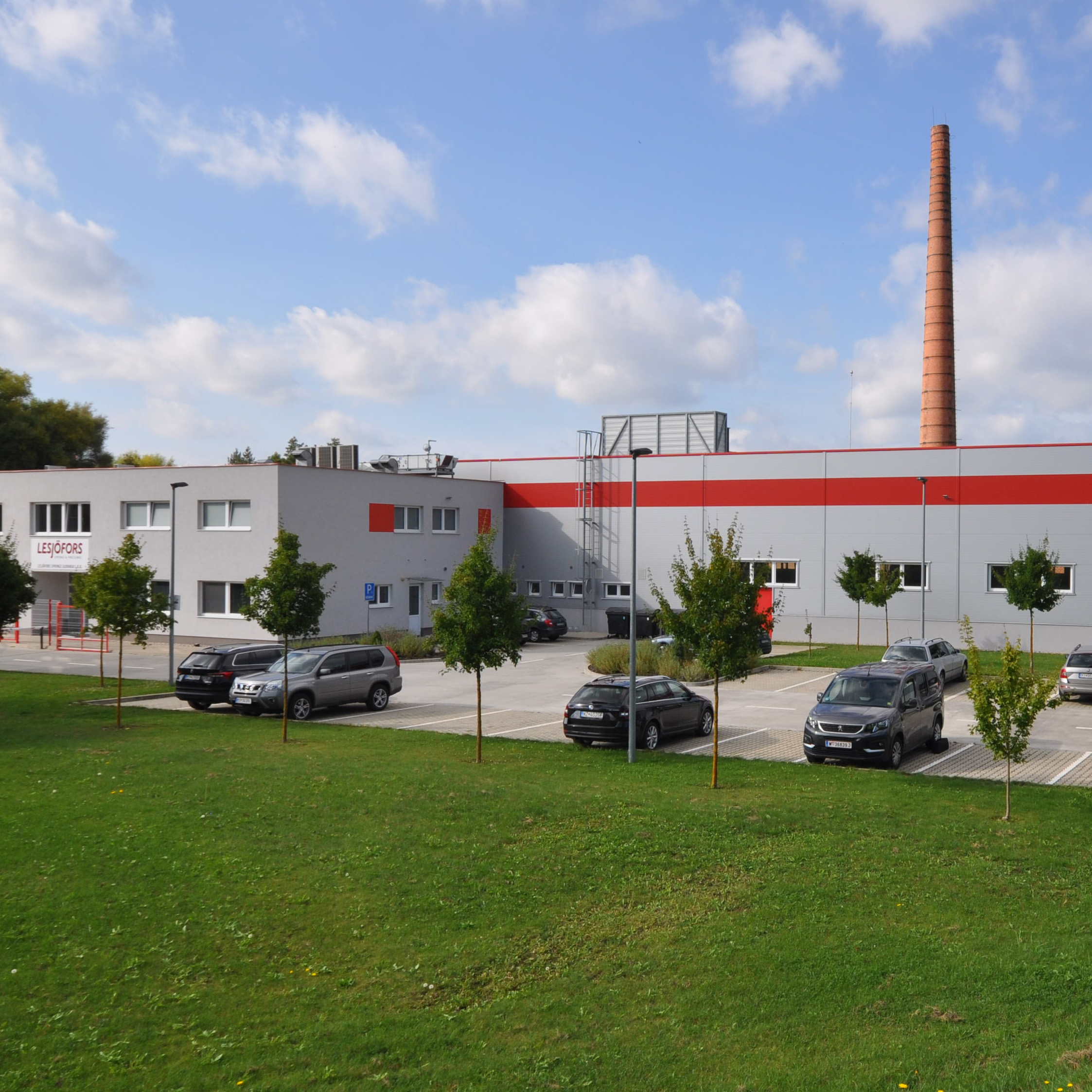 Modern industrial building with red accents, surrounded by greenery and parked cars. A tall chimney rises against a blue sky with scattered clouds.