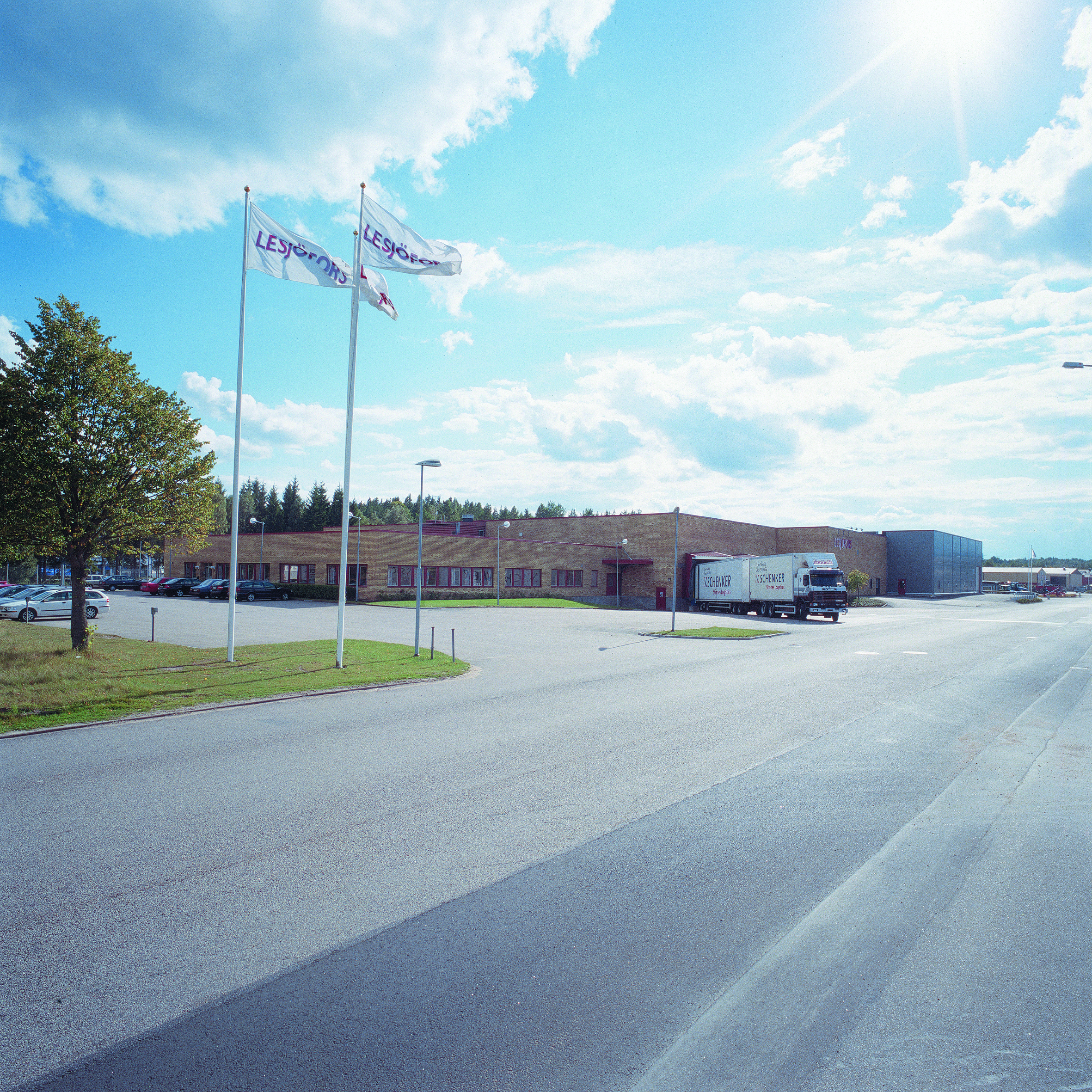 Wide view of an industrial building with brick facade under a partly cloudy sky. Two flags wave in the foreground, and a truck is parked near the entrance.