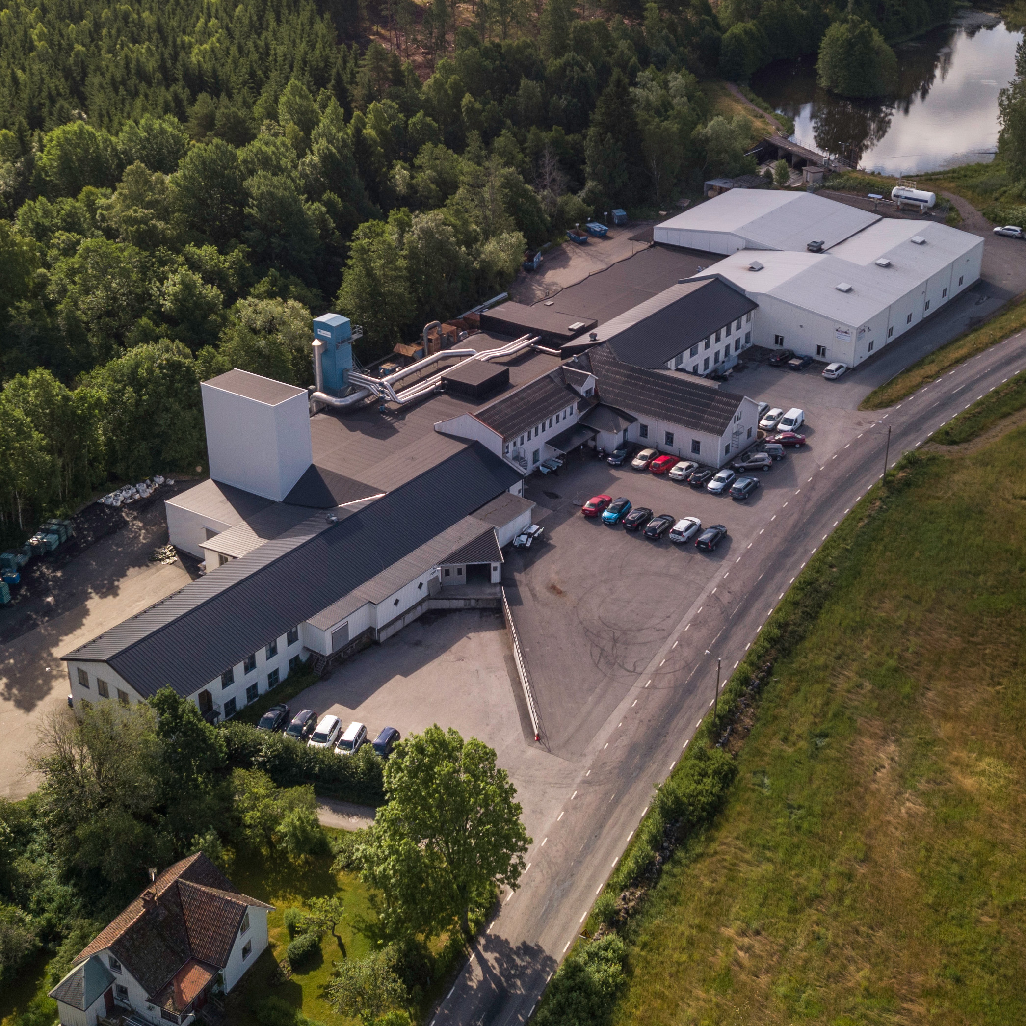 Aerial view of an industrial facility surrounded by lush greenery and trees. Multiple buildings with parked cars are visible, conveying a peaceful, rural setting.