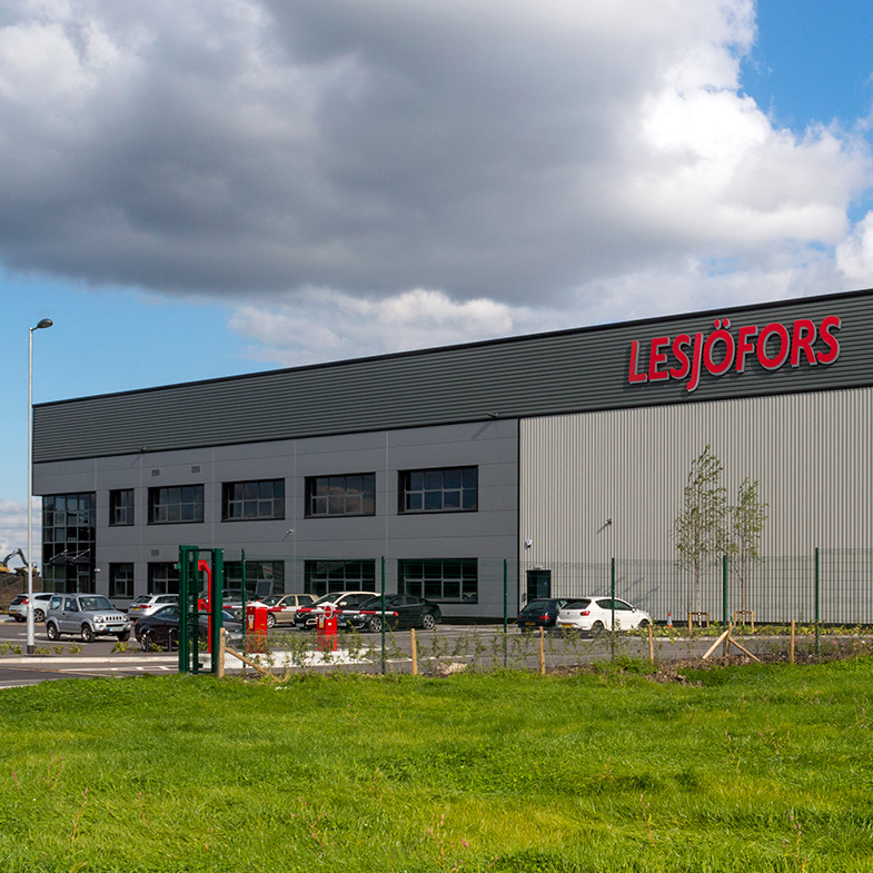 Large industrial building with "Lesjöfors" signage, surrounded by a grassy field under a partly cloudy sky.