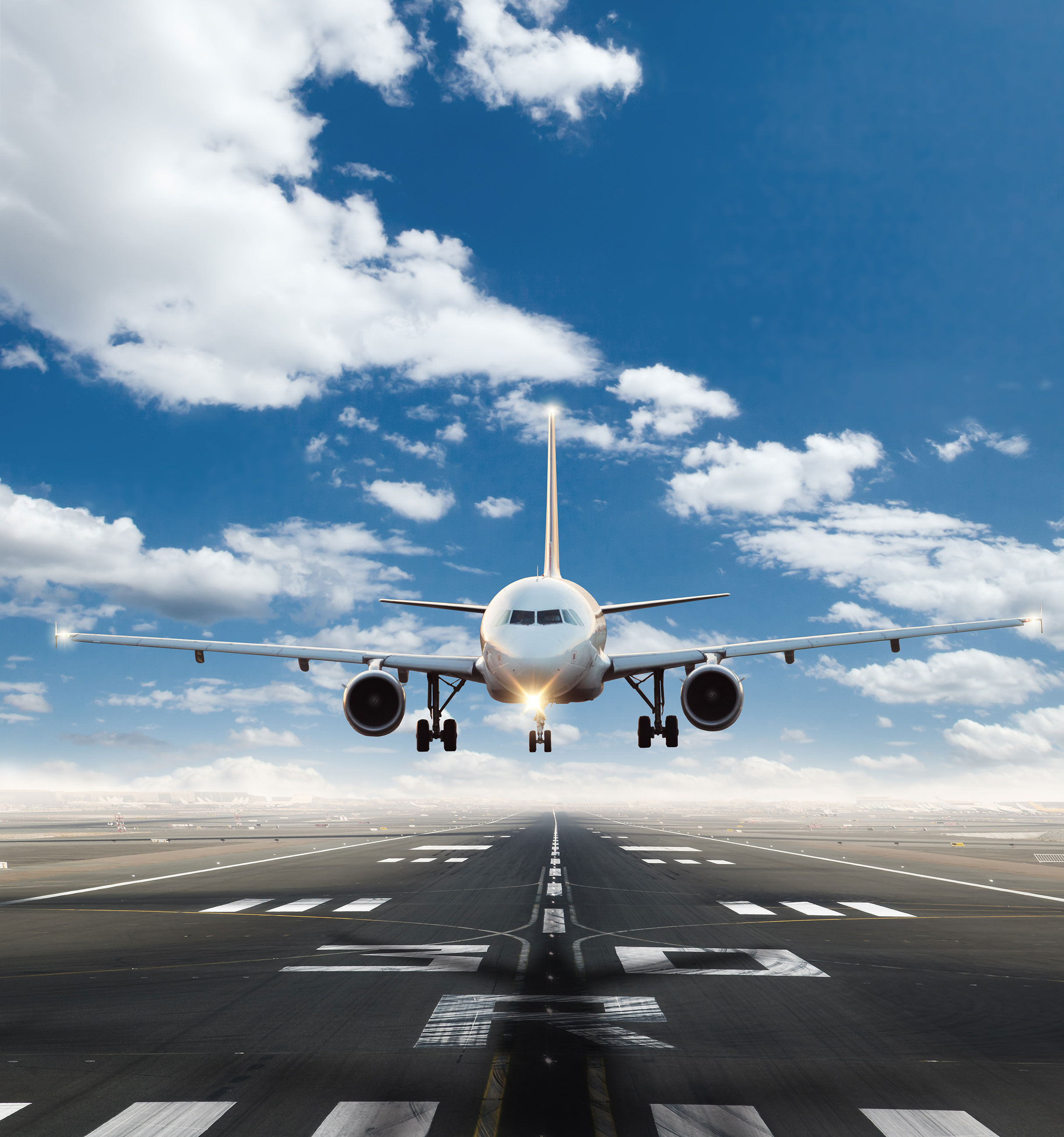 Front view of a white commercial airplane approaching a runway for landing under a blue sky with scattered clouds.