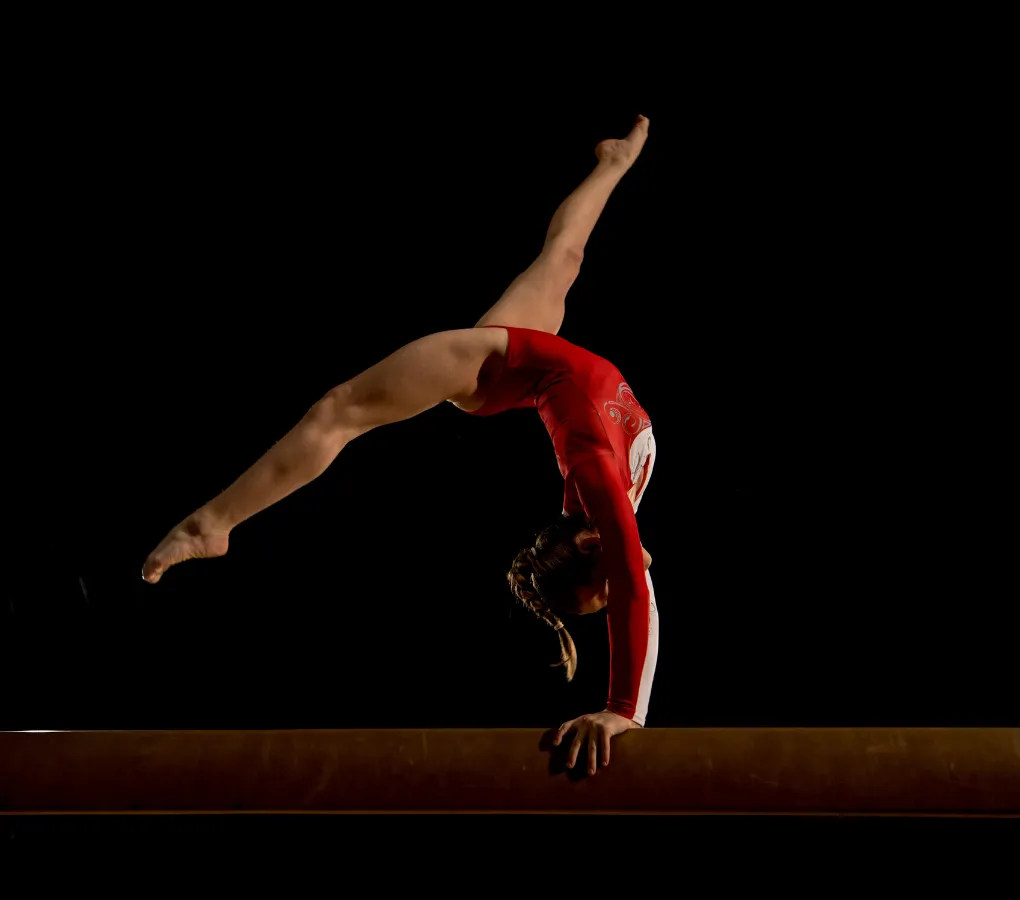 A female gymnast performing a one-armed handstand on the balance beam, wearing a red leotard against a black background