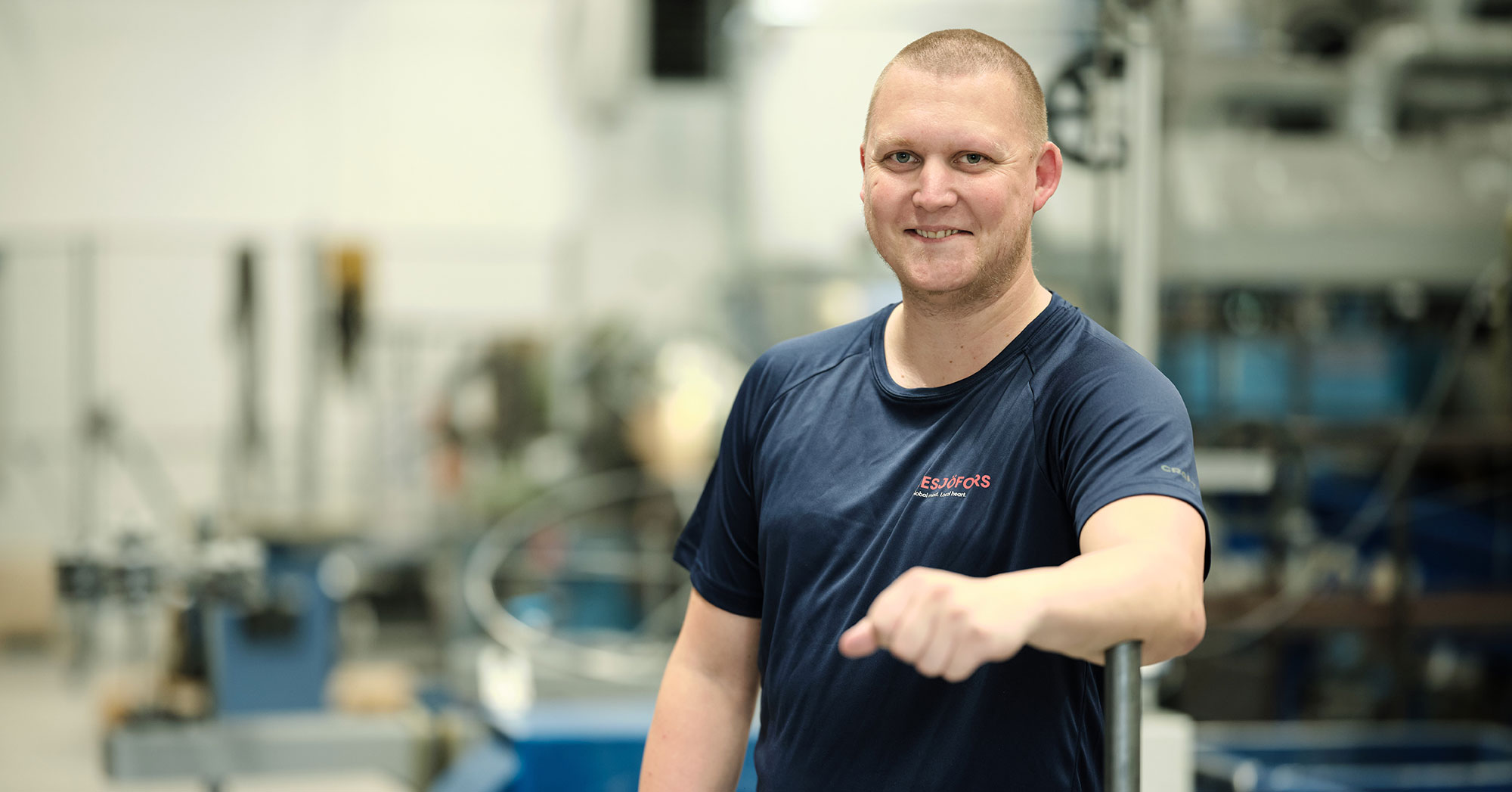 A smiling man in a blue shirt stands confidently in a factory setting, with machinery blurred in the background.