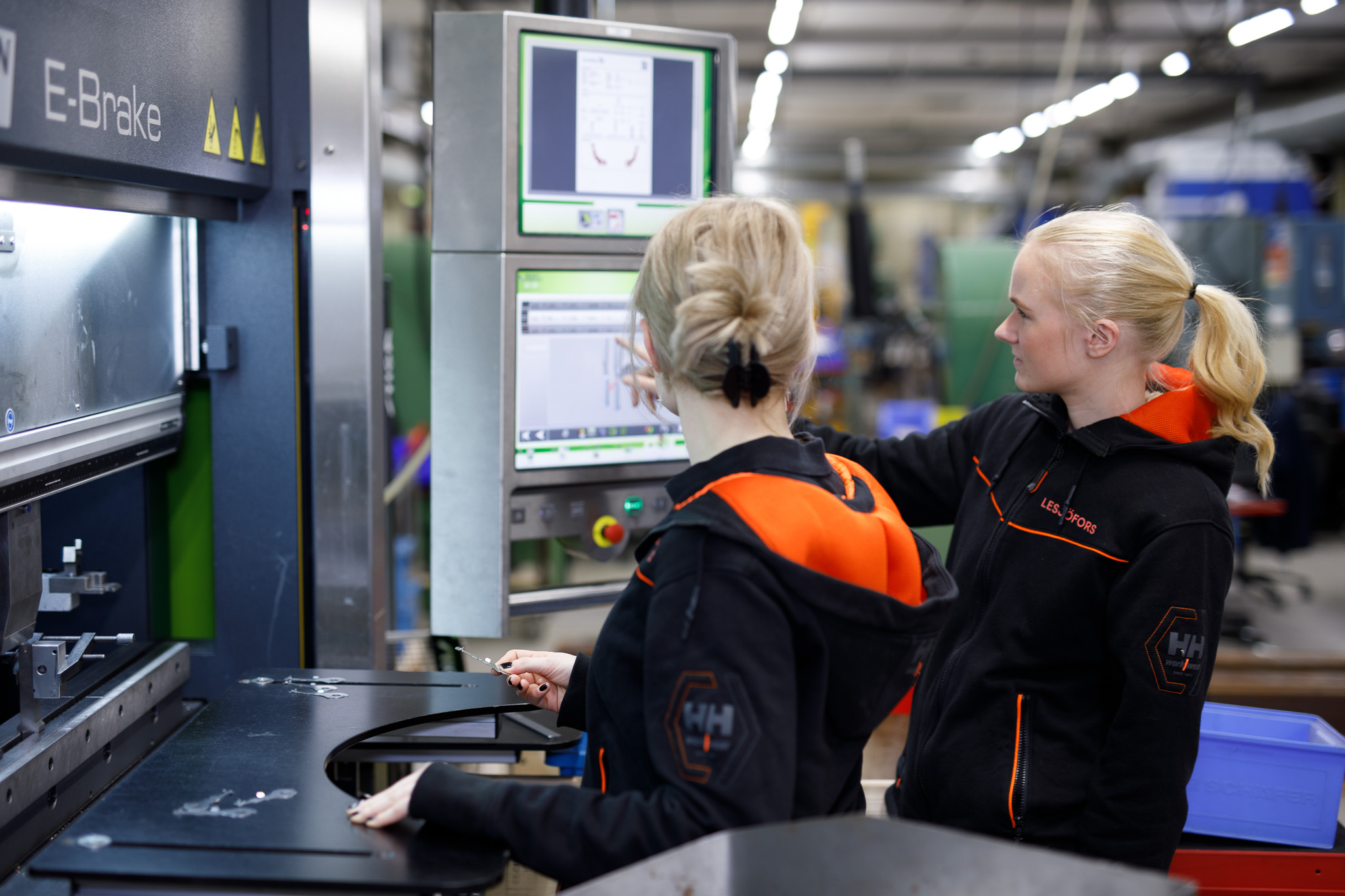Two workers in a factory, wearing black and orange uniforms, operate machinery with digital screens.