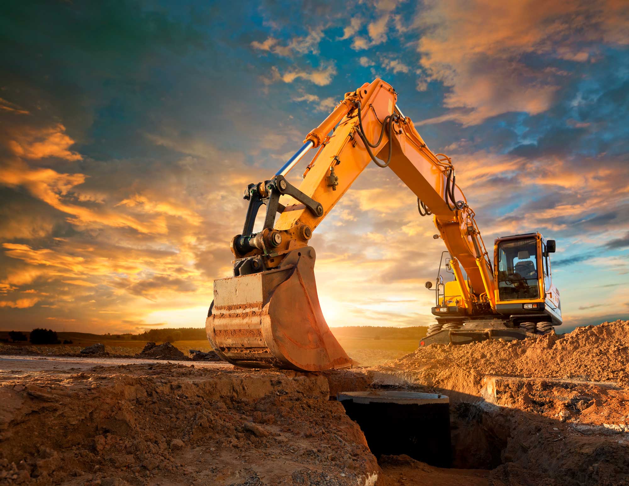 A large excavator at a construction site is silhouetted against a dramatic sunset sky, with vibrant orange and blue hues.