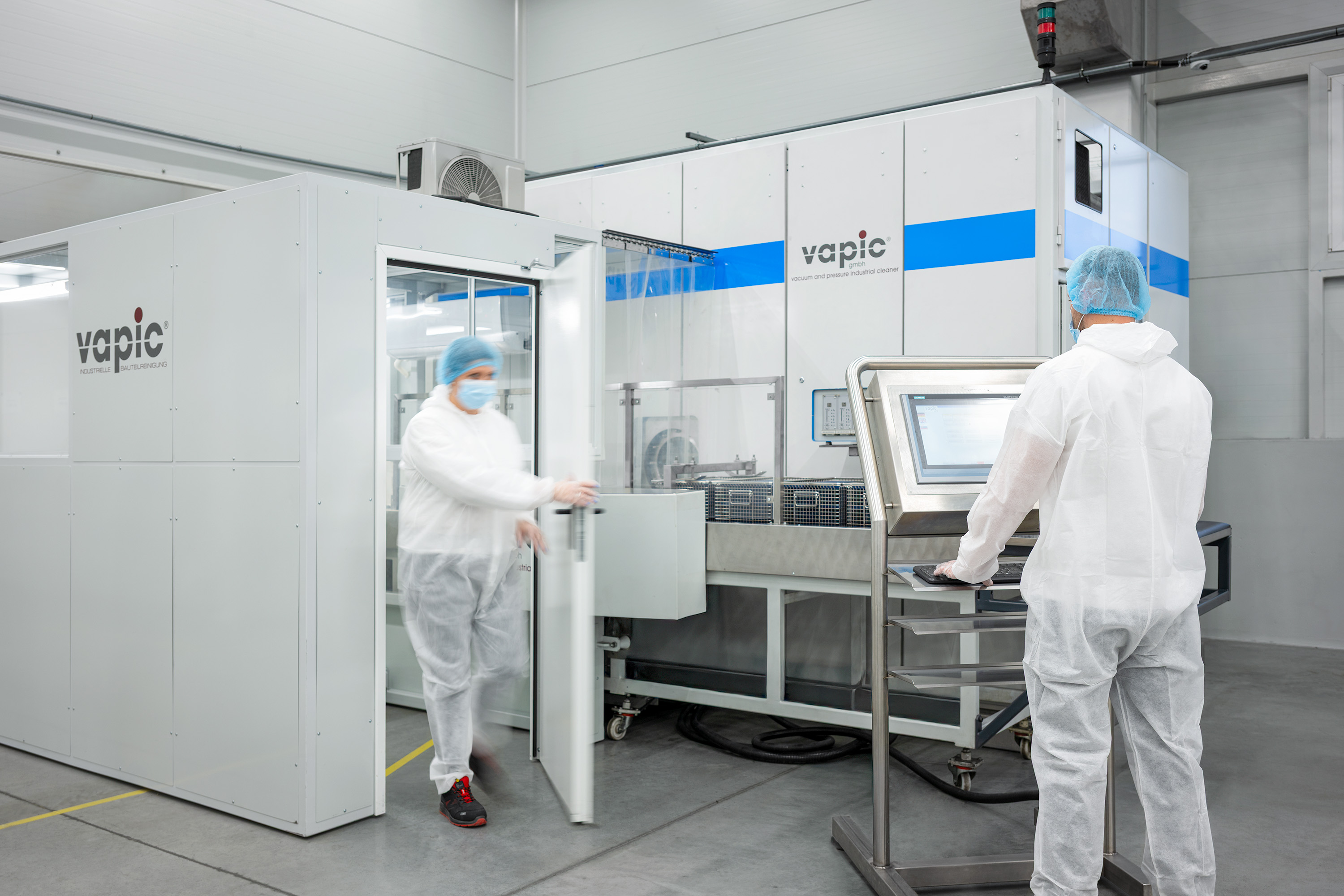 Technicians in white protective suits operate machinery in a sterile, modern laboratory.