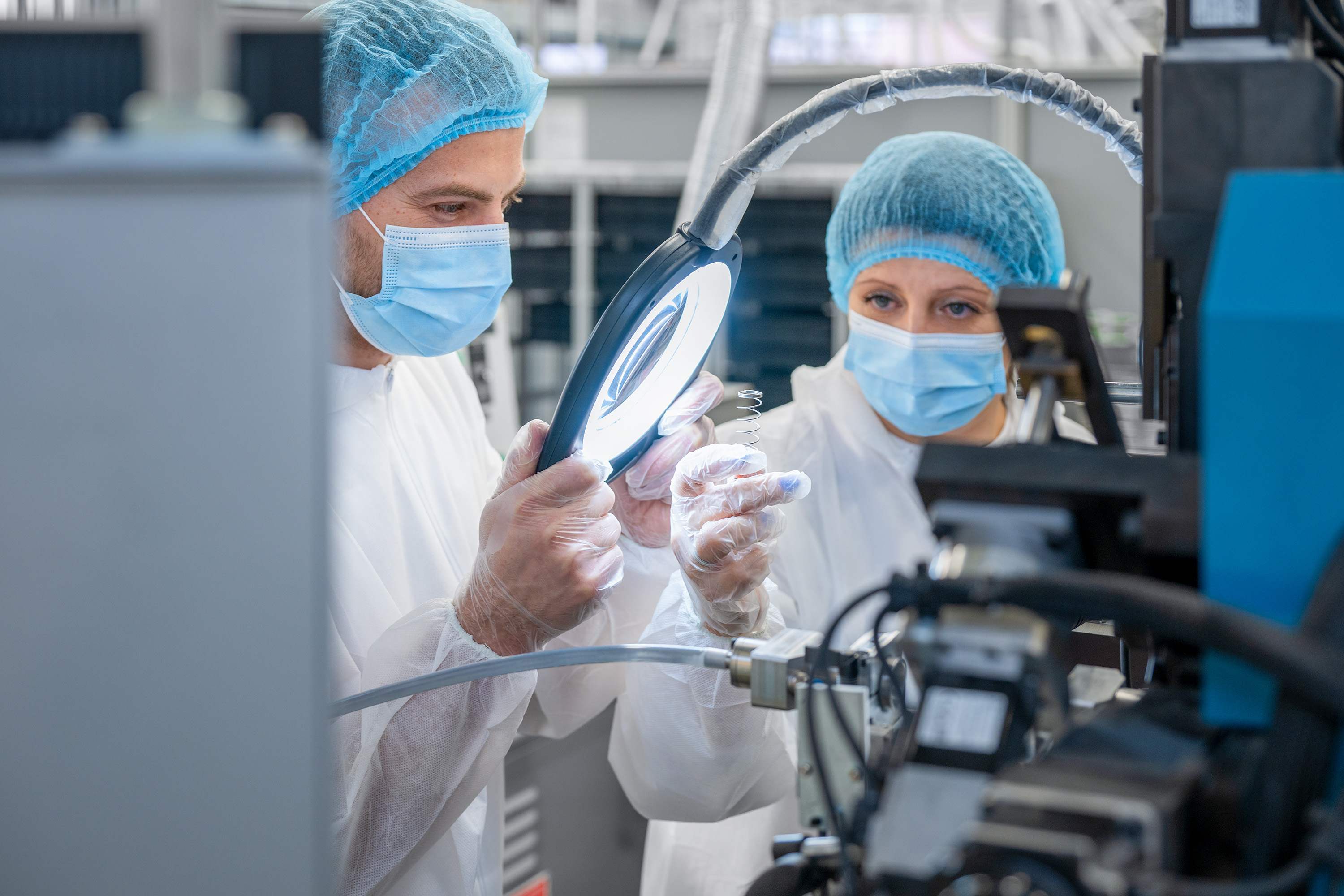 Two scientists in protective gear and masks work in a lab, examining a small object under a magnifying glass. The setting is technical and focused.