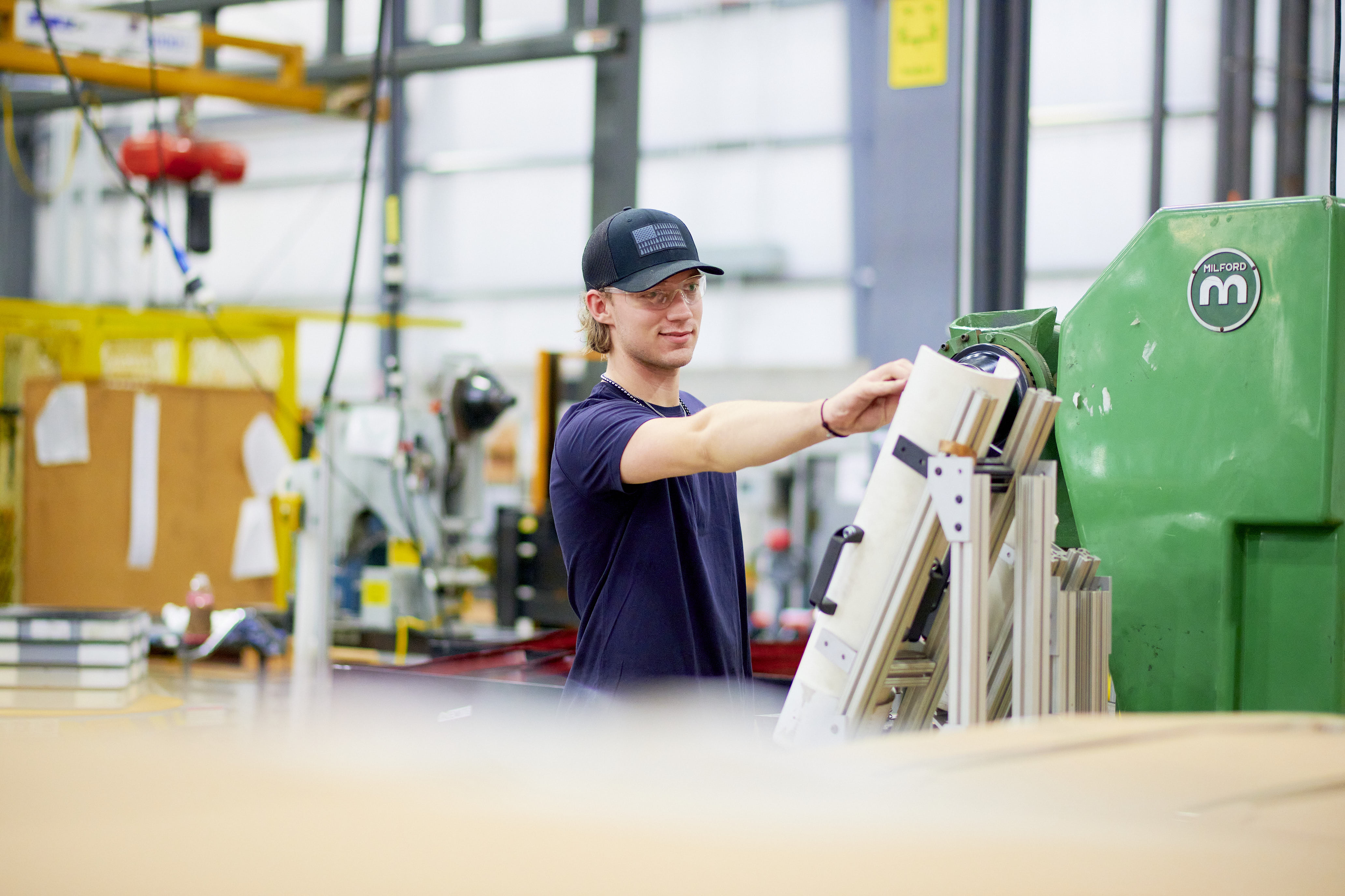 A worker in a cap operates machinery in a bright, spacious factory. The scene conveys focus and industrial efficiency with modern equipment visible.