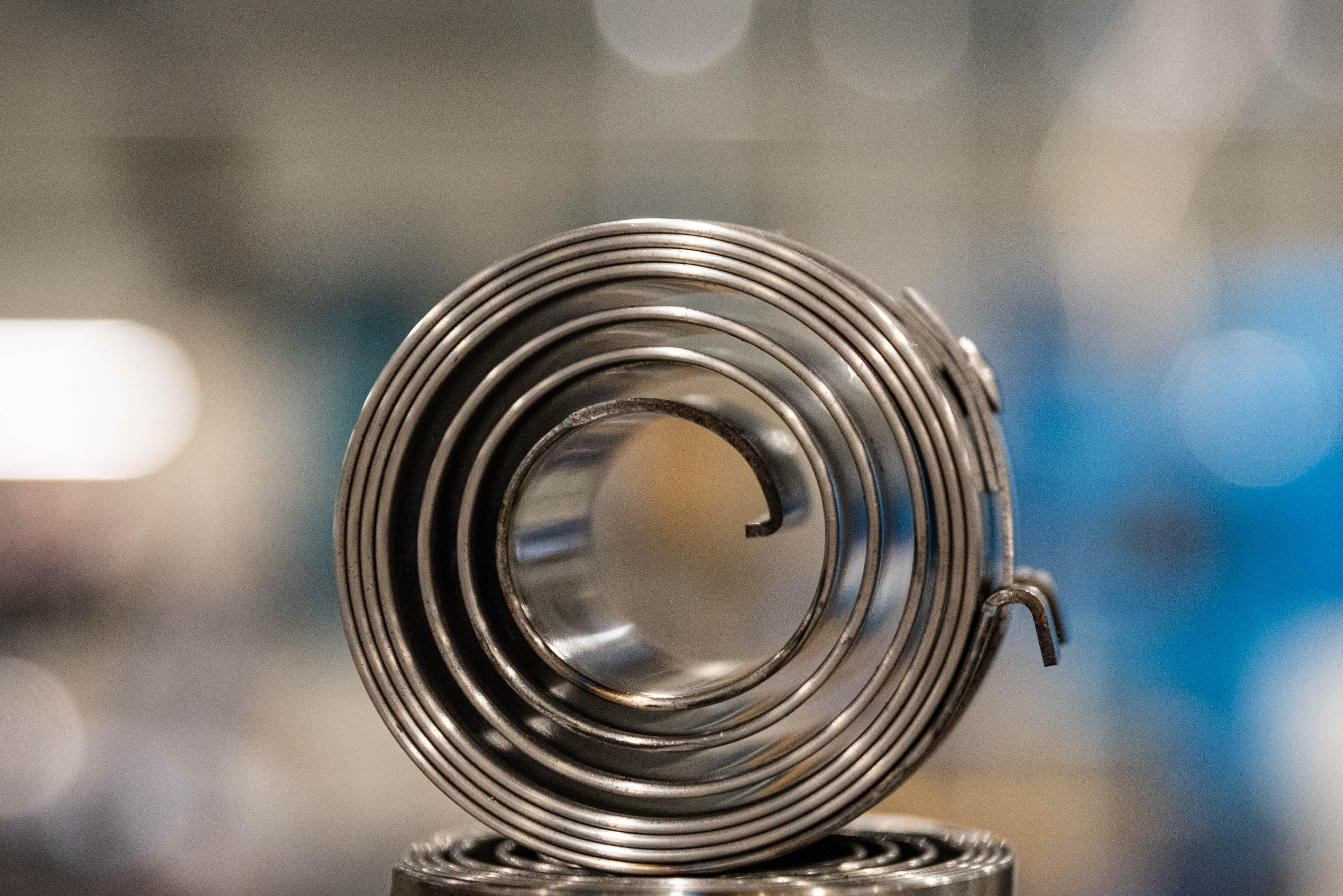 Close-up of a metallic power spring in a workshop setting, with a blurred background. The spring's shiny coils evoke precision and industrial craftsmanship.