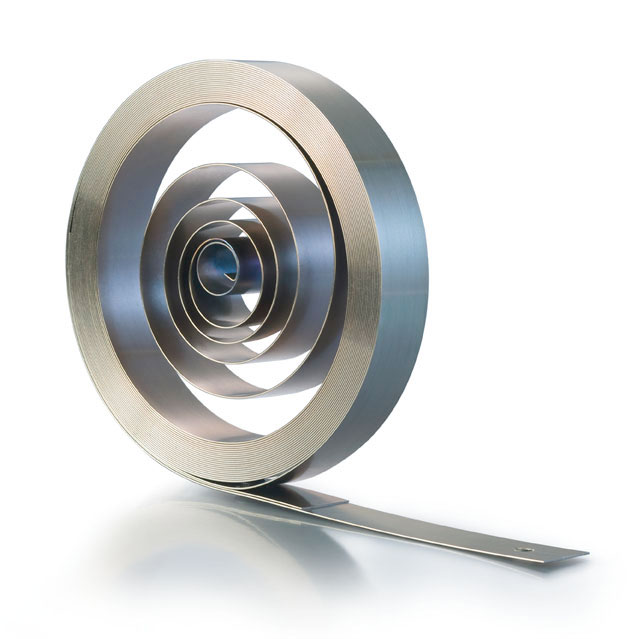 A neatly coiled power spring of flexible metal sits on a reflective surface. The spiral design creates a sense of balance and precision.