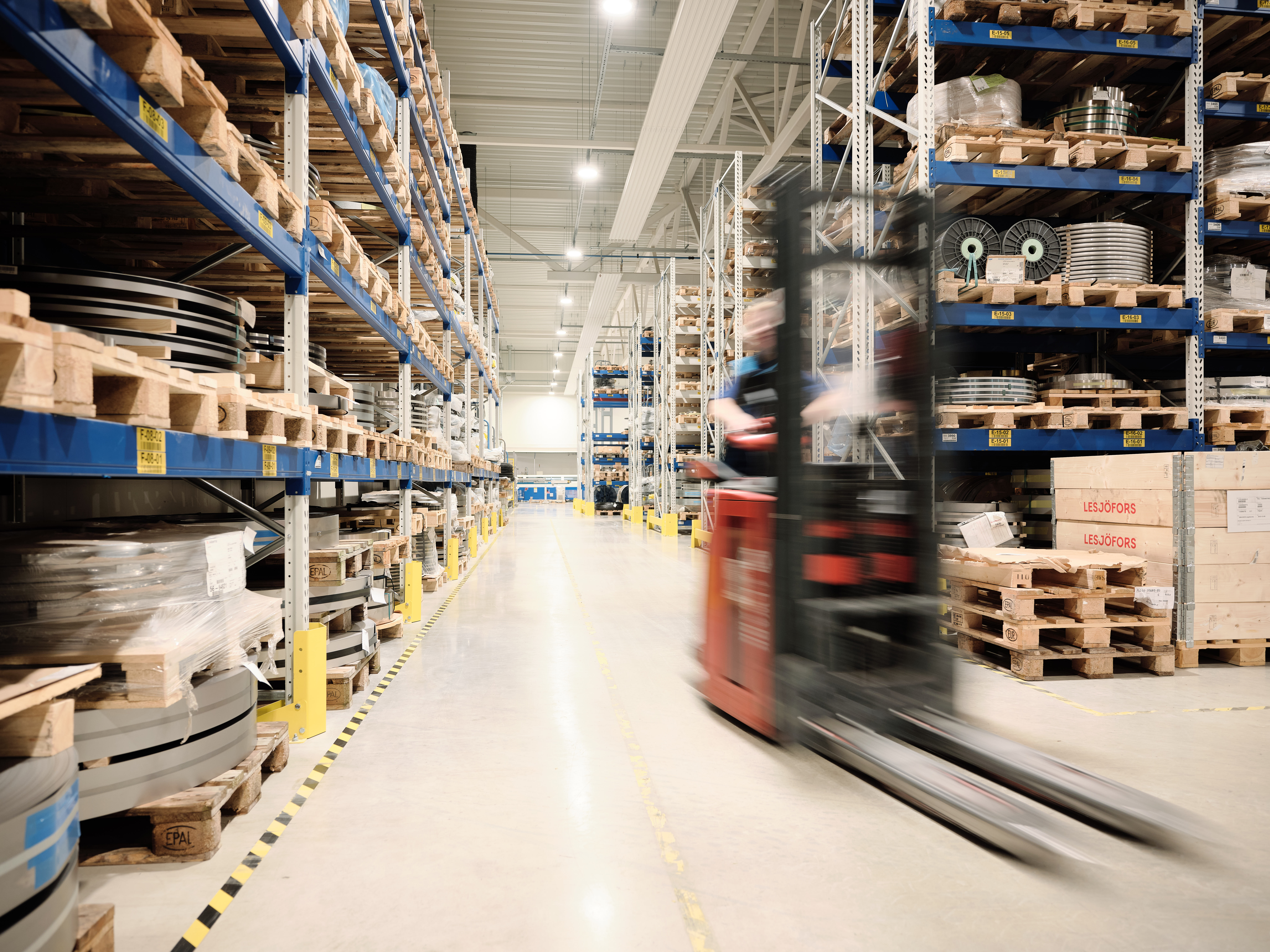 A warehouse scene with tall shelves full of boxes. A forklift moves quickly down the aisle.