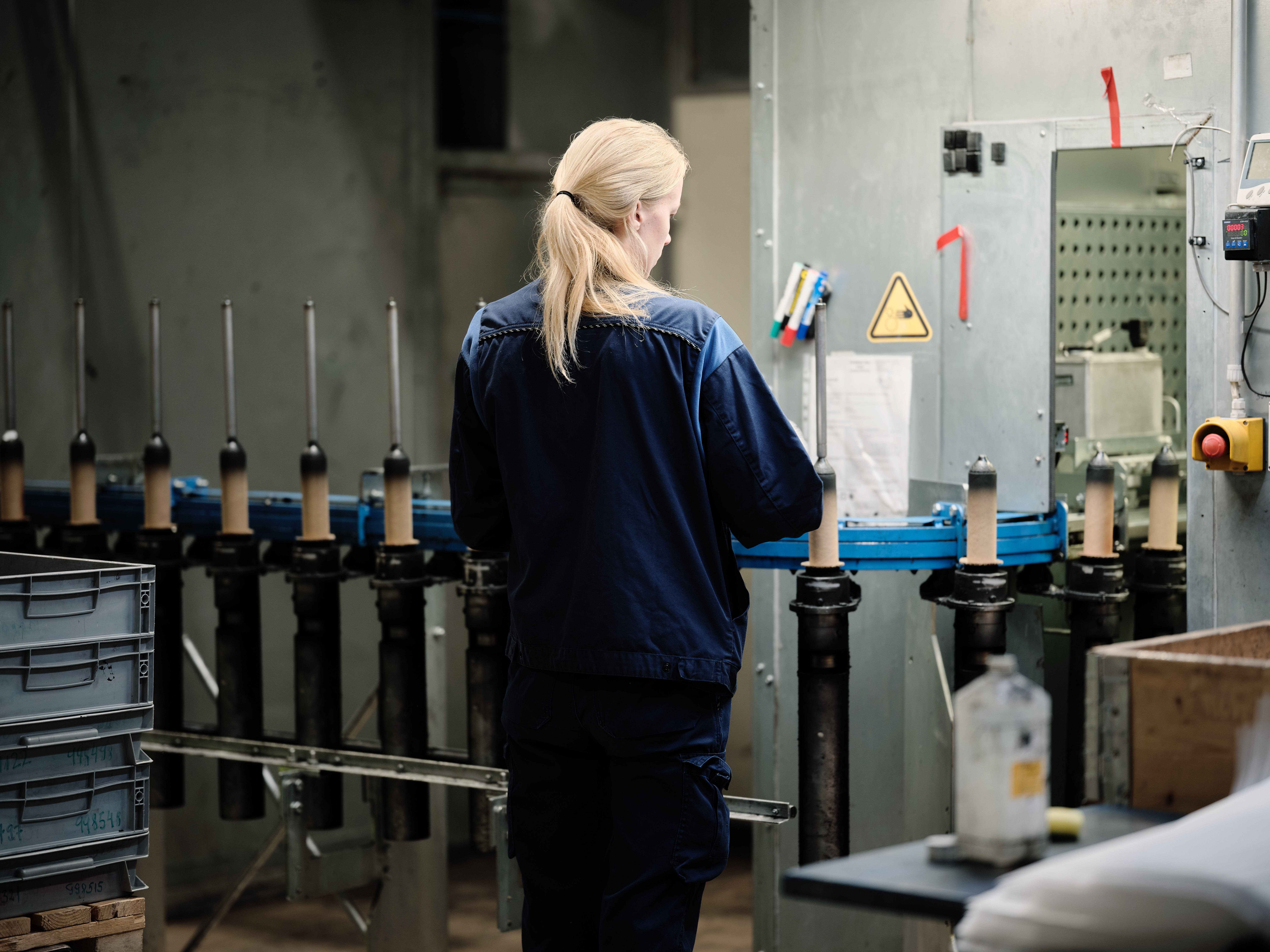 A person with long blonde hair, wearing a dark blue work uniform, stands with their back to the camera and operates machinery in an industrial workshop with lockable gas springs. Rows of metal components and equipment are visible on workbenches around them.