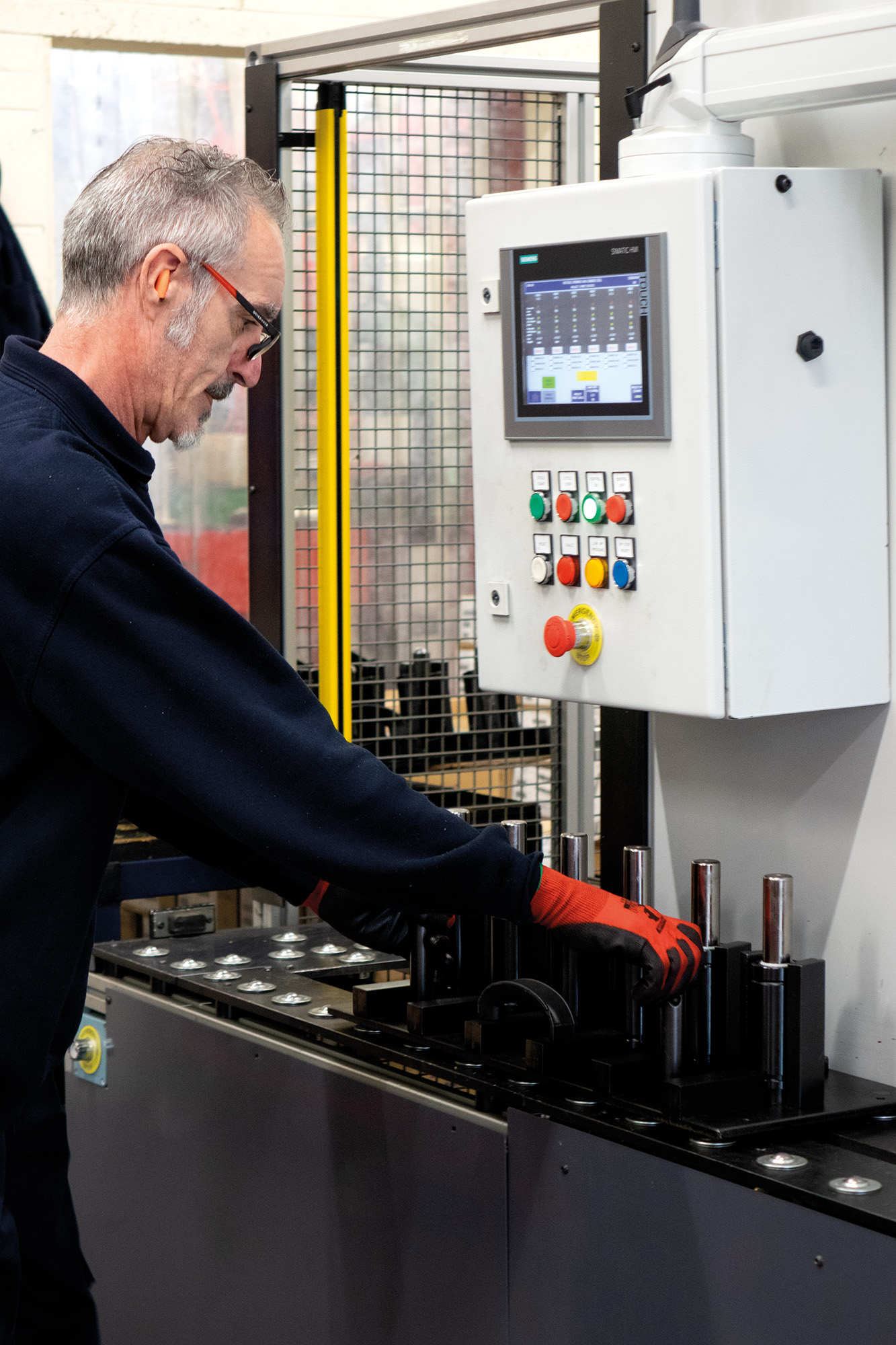 A person with gray hair operates industrial machinery, pressing controls. They wear safety gear, including red gloves. A digital display is visible.