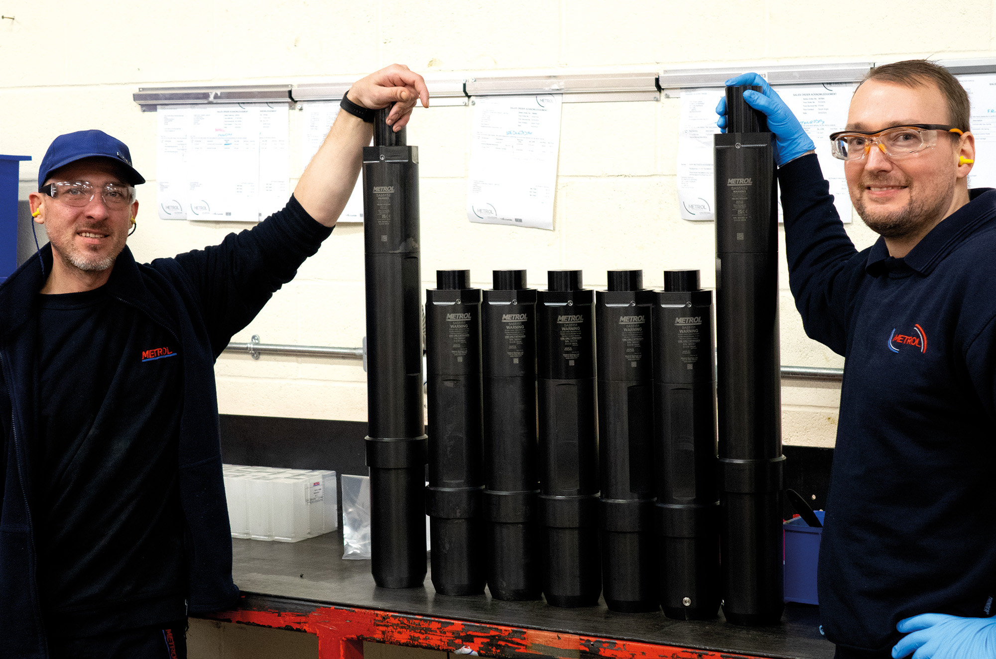 Two people in a workshop, smiling and holding large black cylindrical objects. They stand on either side of a table, with several cylinders in between.