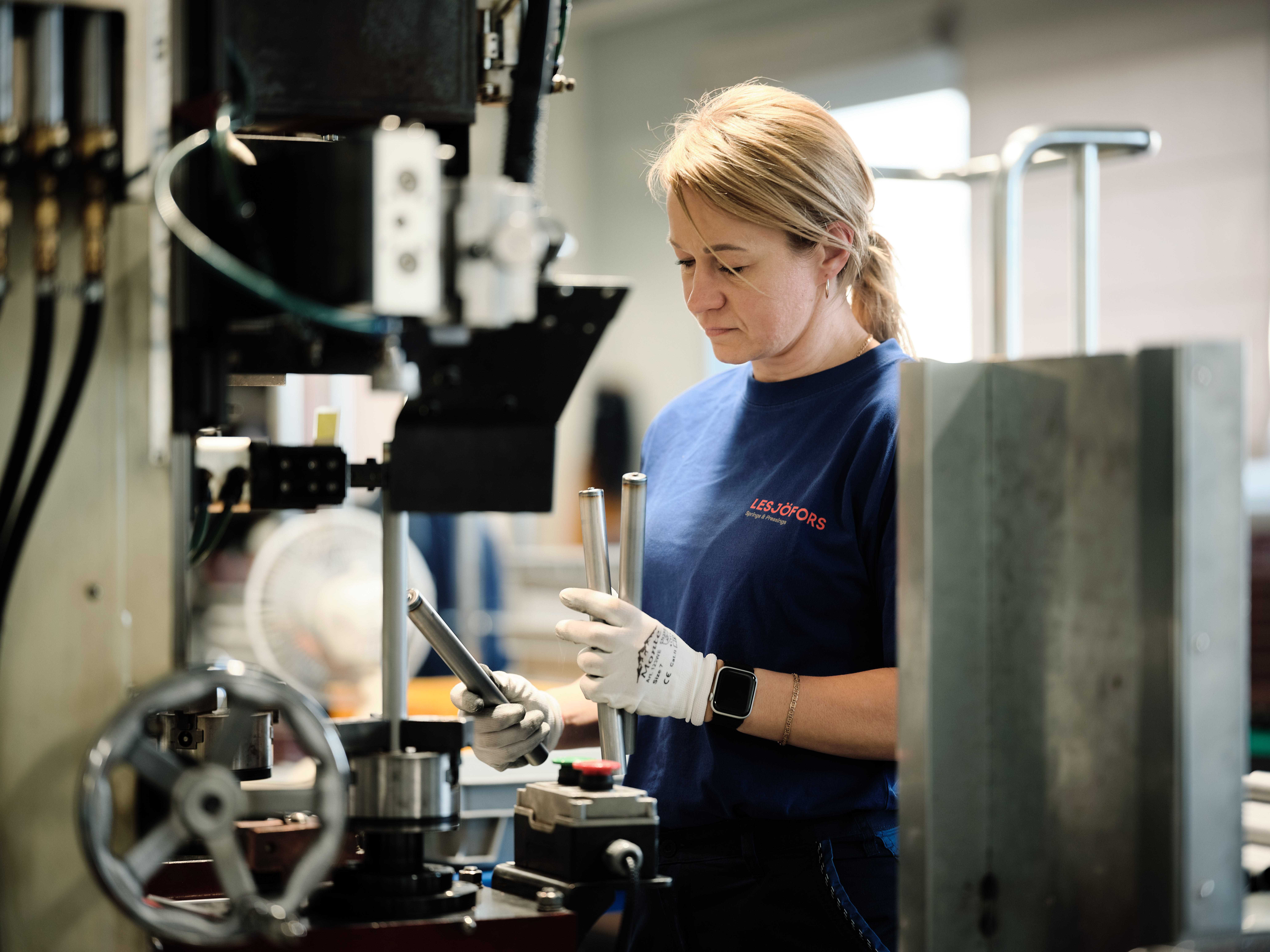 A woman in a factory setting, wearing a blue shirt and gloves, focuses on operating machinery.