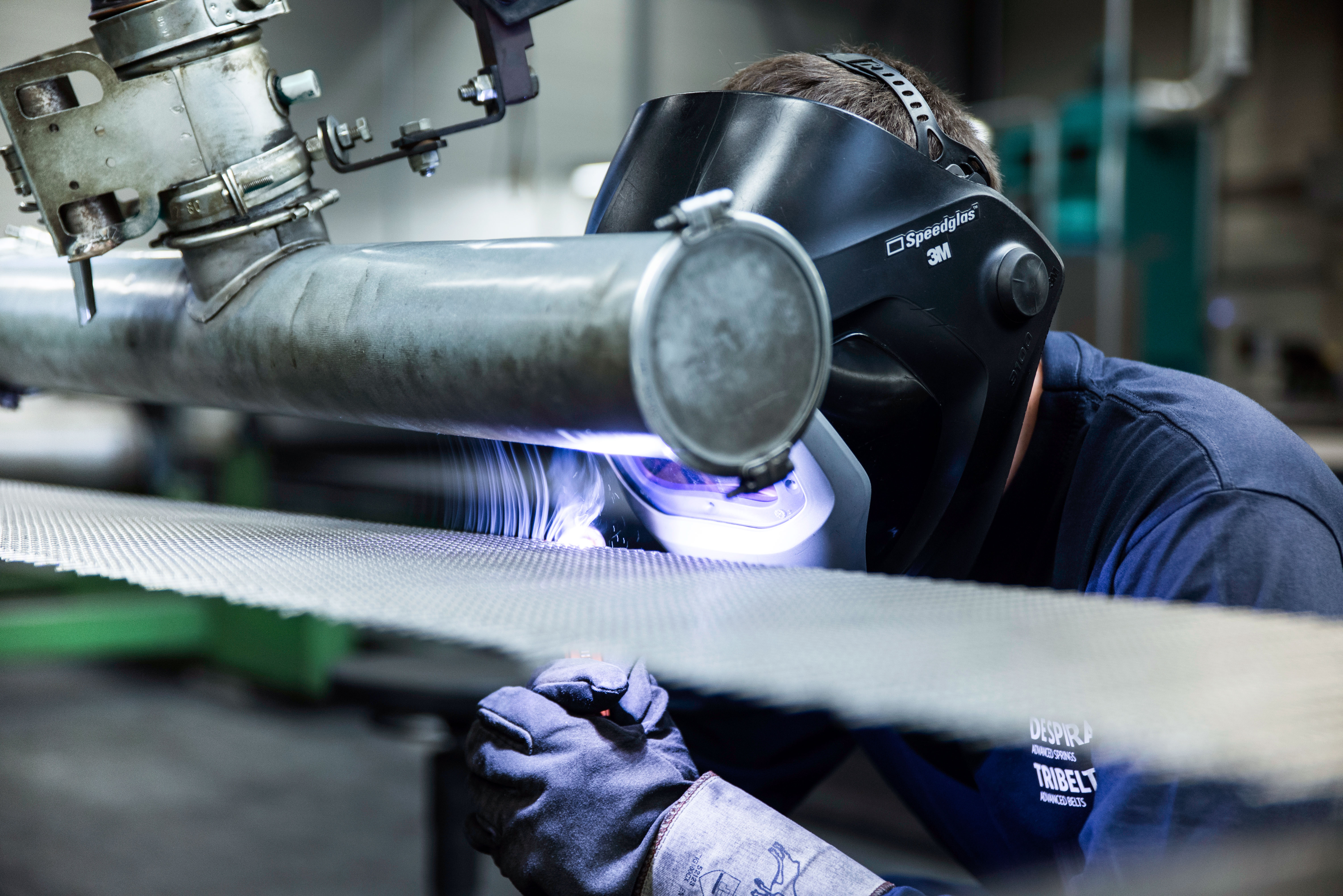 Worker in a factory wearing a welding helmet and gloves, focused on welding a metal pipe. Sparks fly, highlighting precision and concentration.