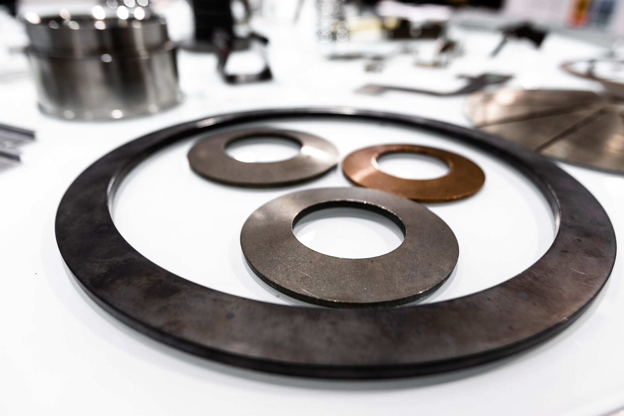 A large metal disc spring displayed on a white table, with three smaller disc springs of different materials and diameters arranged inside it.