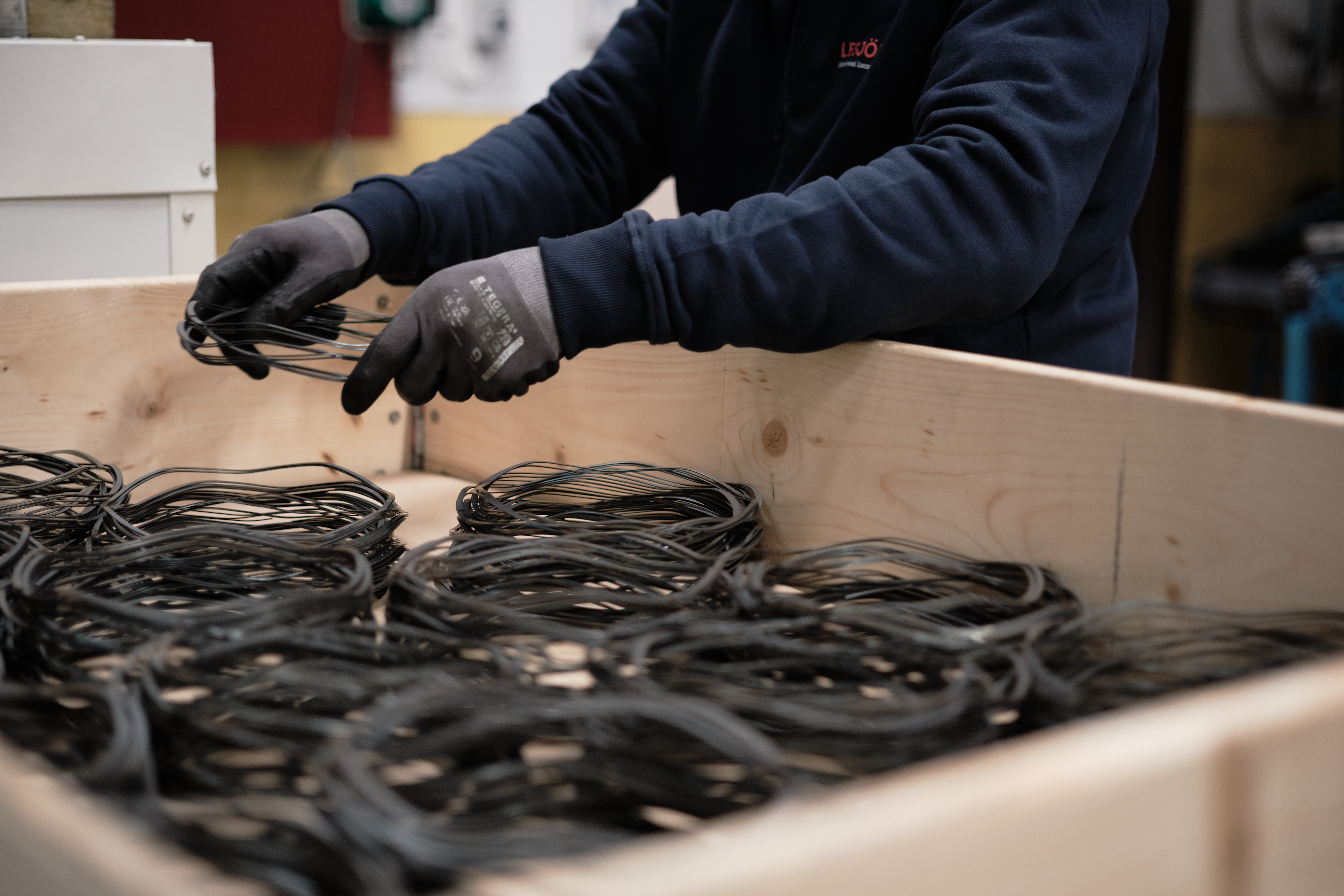Person in gloves sorting wave springs in a large wooden box. The setting appears industrial.