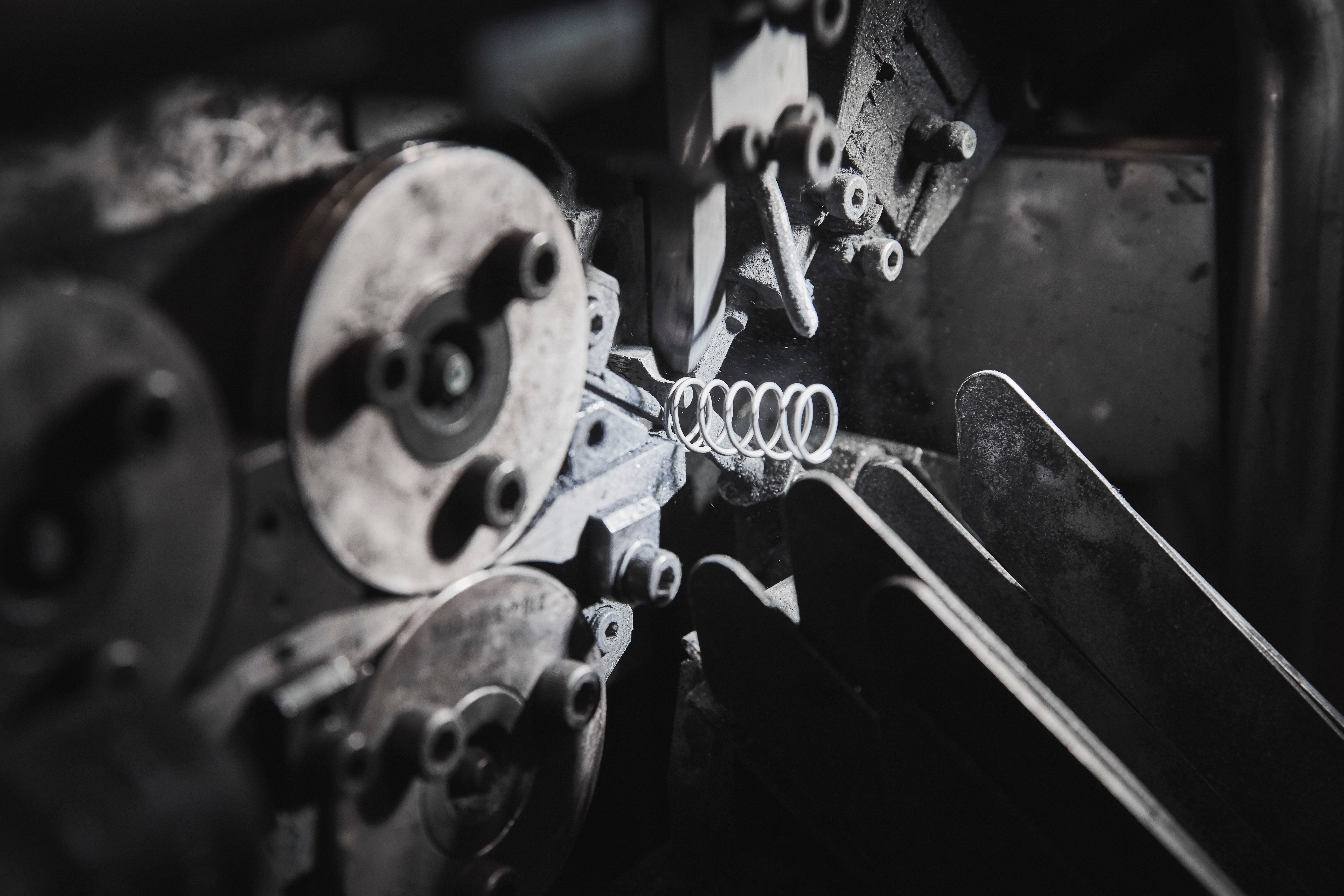 Close-up of a metal coil spring being formed inside a spring coiling machine, with wire feeding through rollers under low-light conditions.